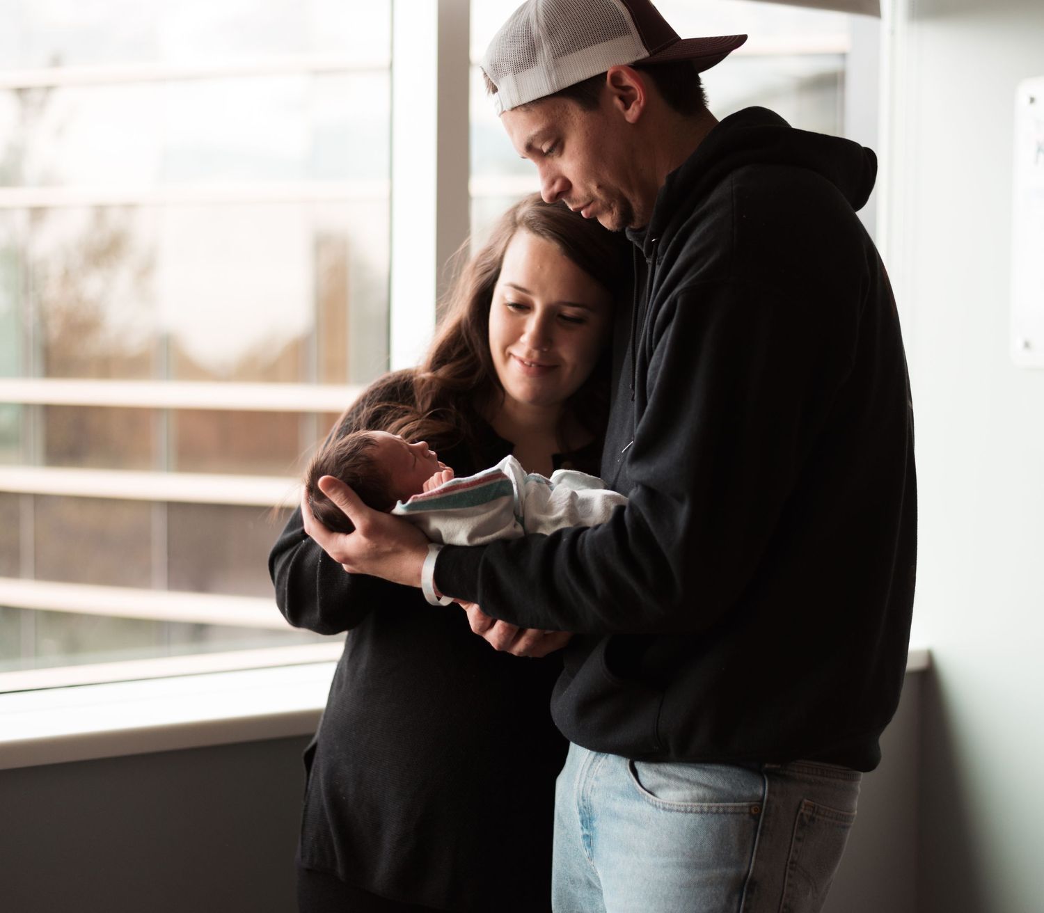 Black and white portrait series showing tender moments between parents and newborn by a window.
