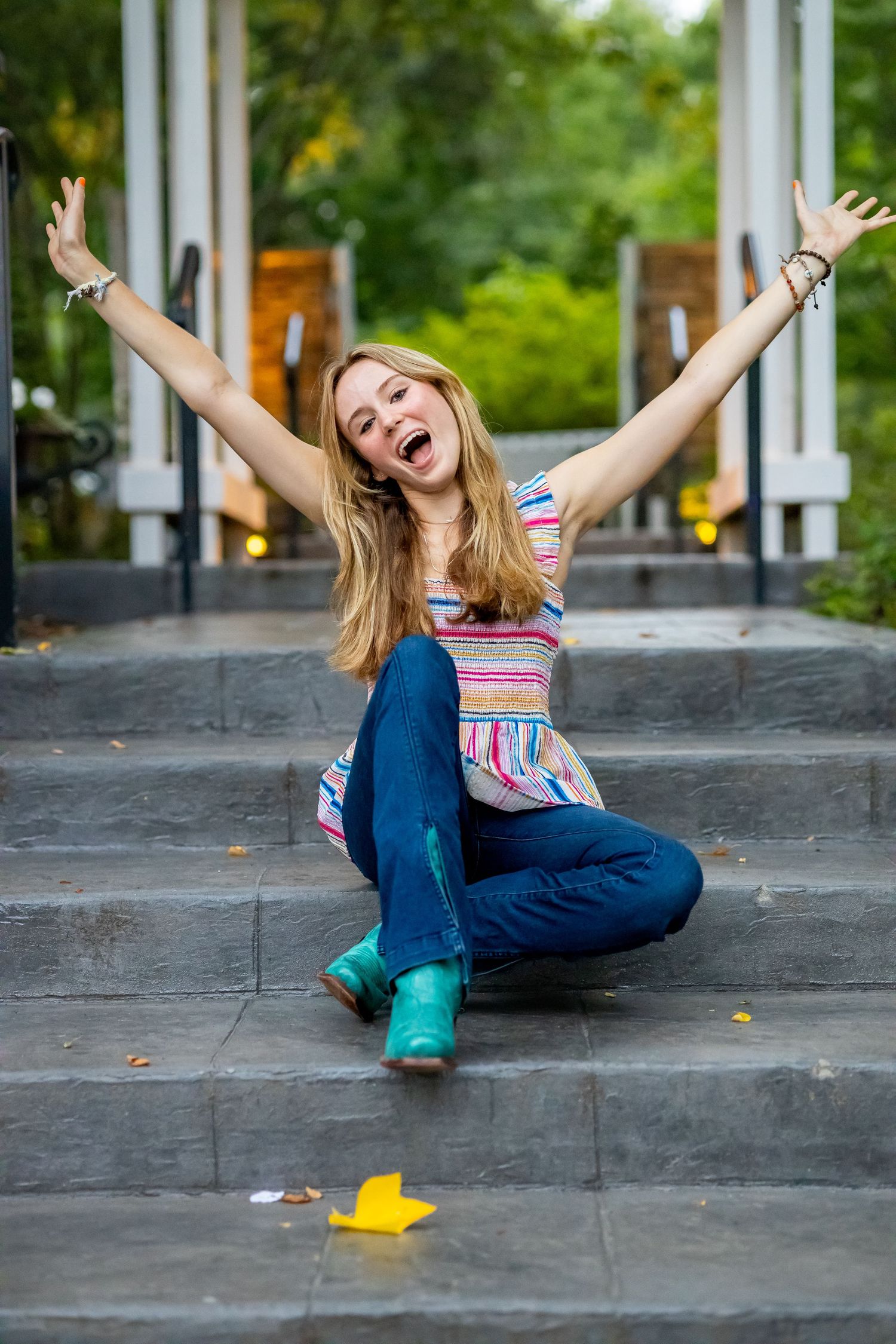 Joyful portrait on concrete steps with arms raised wearing striped top and turquoise boots.