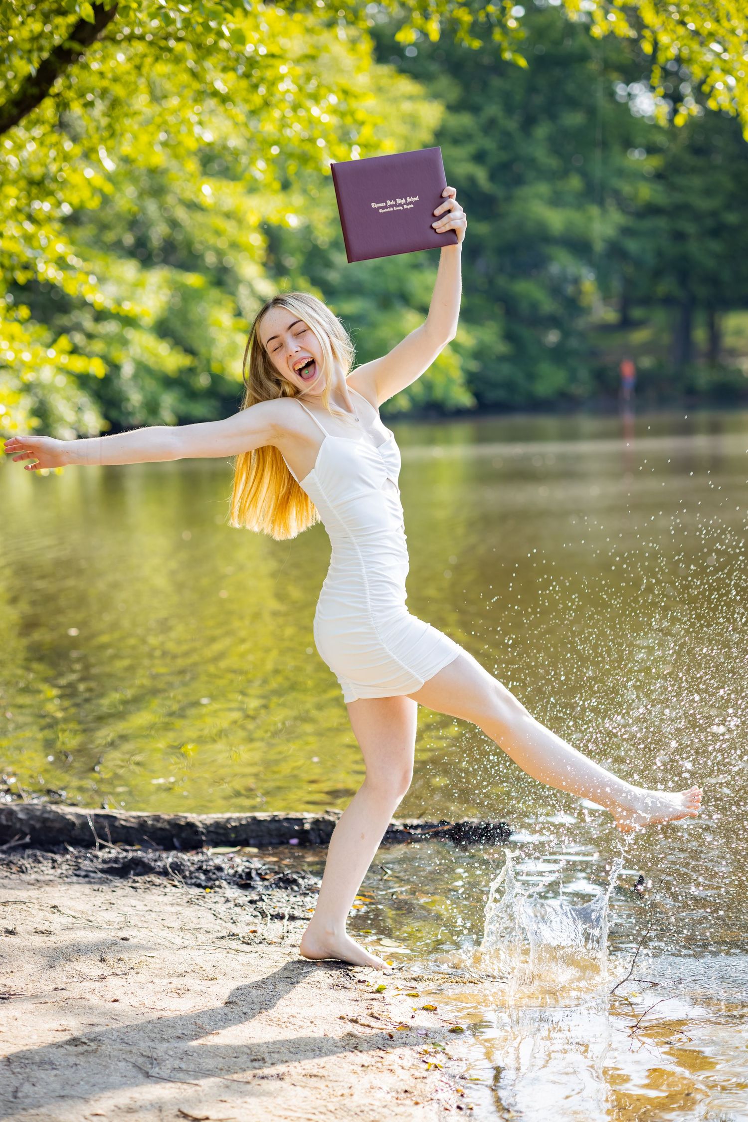 Excited graduate splashes in water while celebrating with diploma by lake.
