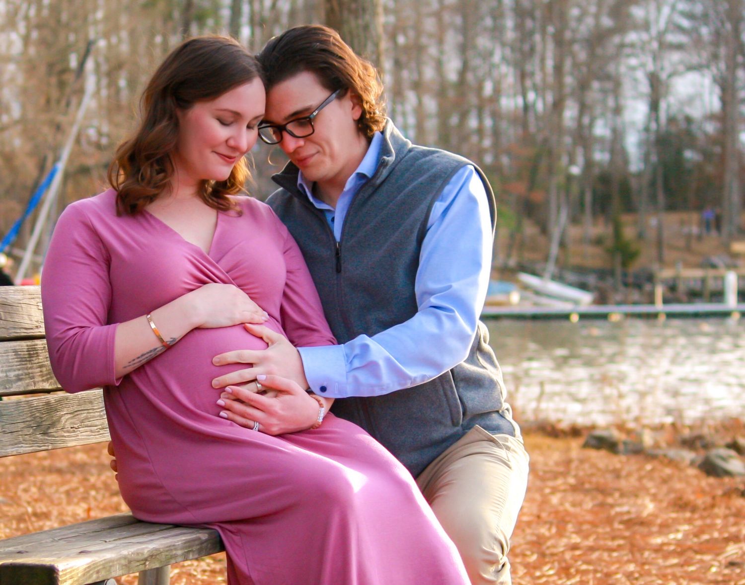 Expecting couple shares tender moment on bench during maternity photo session by lake.