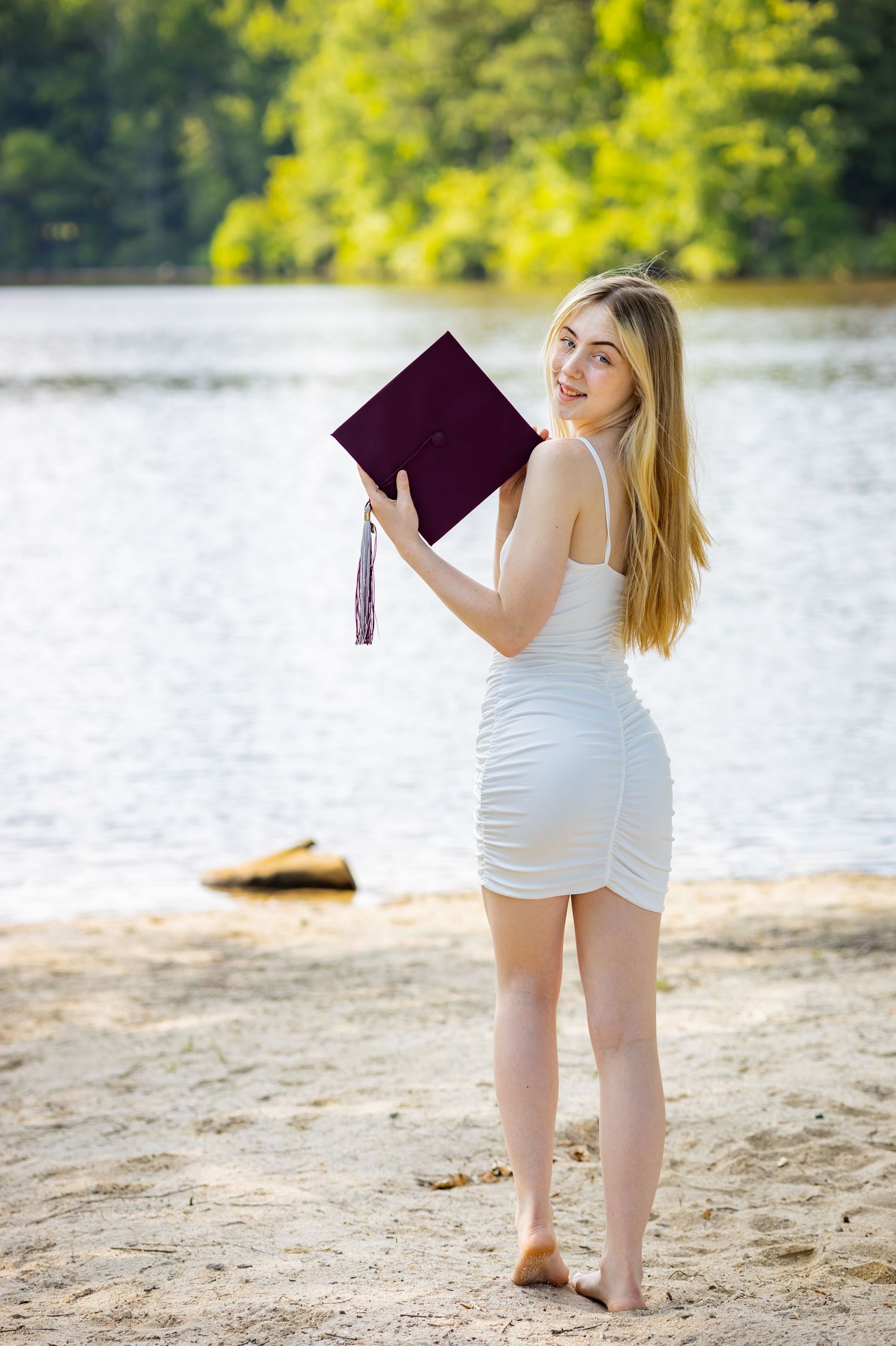 Graduate in white dress holds burgundy diploma on lakeside beach during sunny summer photoshoot.
