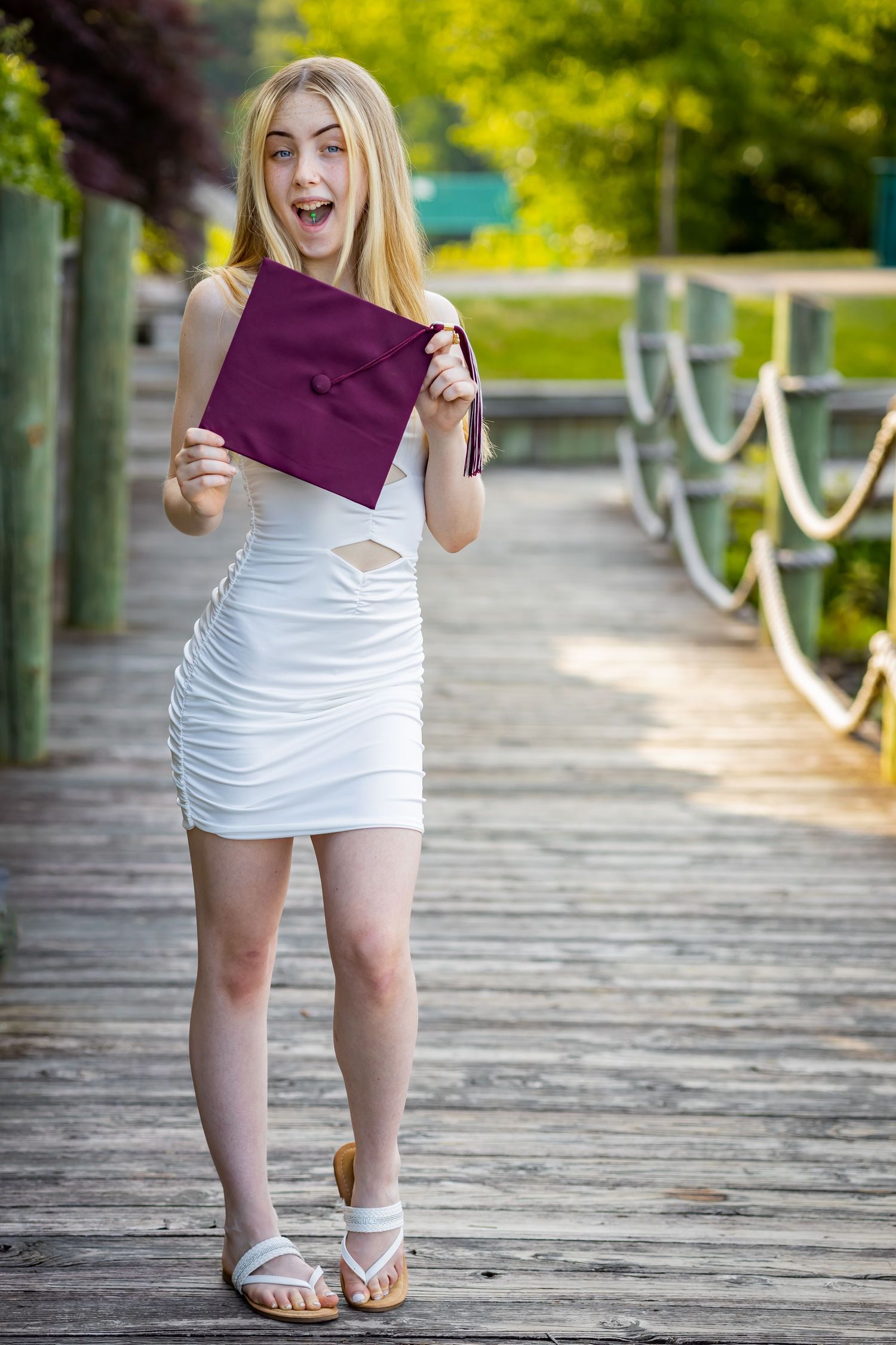 Excited graduate holding maroon diploma cover on wooden boardwalk during celebration.