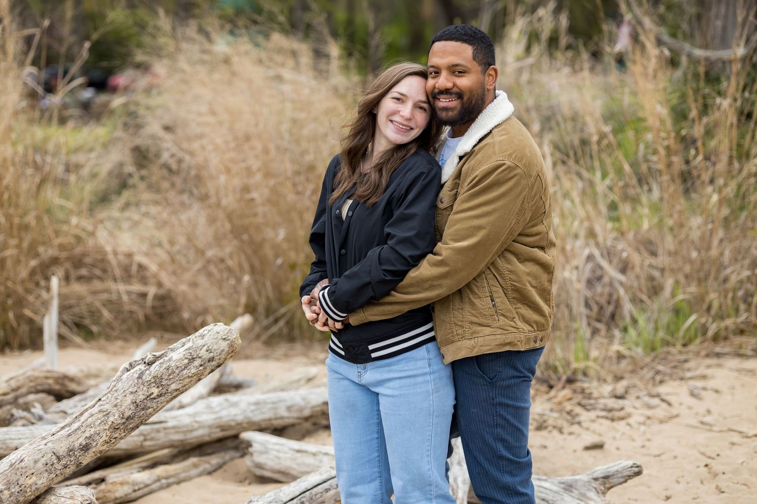 A couple embraces on a beach with dried grass and driftwood in the background.