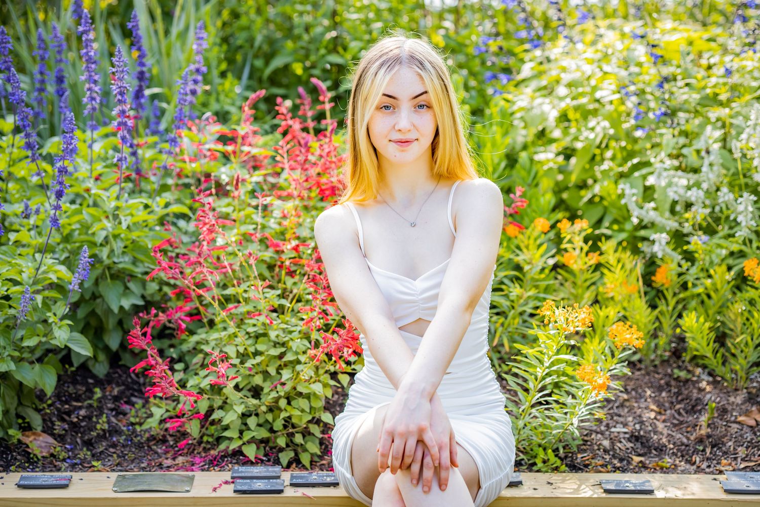 Graduate poses on bench in front of vibrant garden with purple and coral flowers.