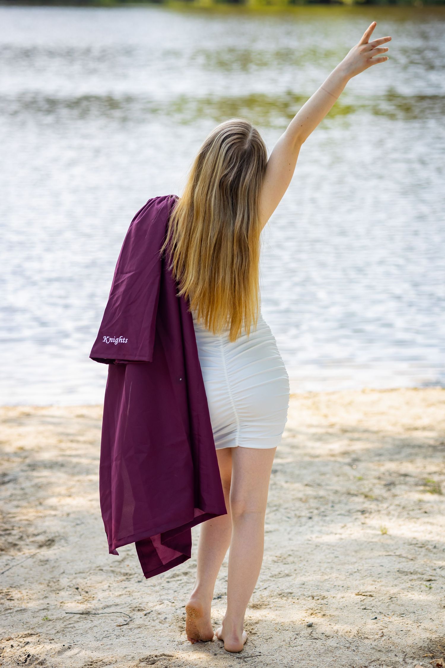Graduate in white dress and maroon gown raises arm triumphantly by waterfront.