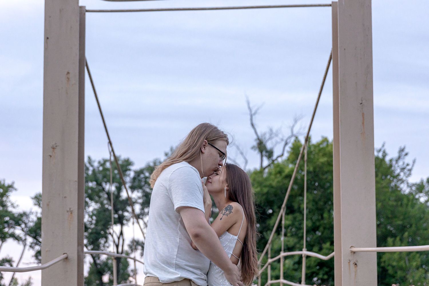 Two people embrace tenderly on a wooden platform outdoors with trees in the background.