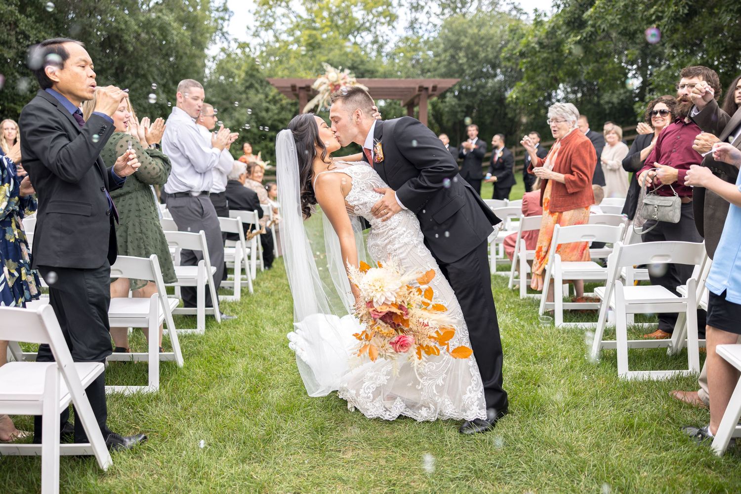 A couple shares their first kiss as newlyweds during an outdoor garden wedding ceremony while guests look on and applaud.
