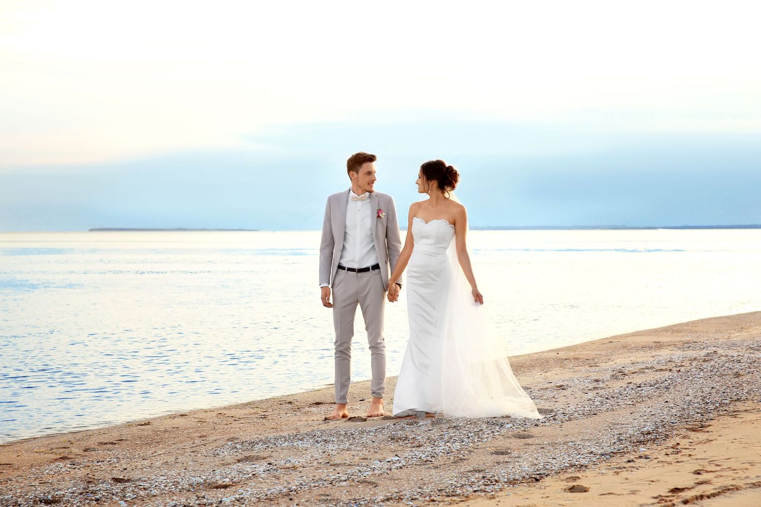 Newlyweds walking along beach shoreline at sunset in formal wedding attire.