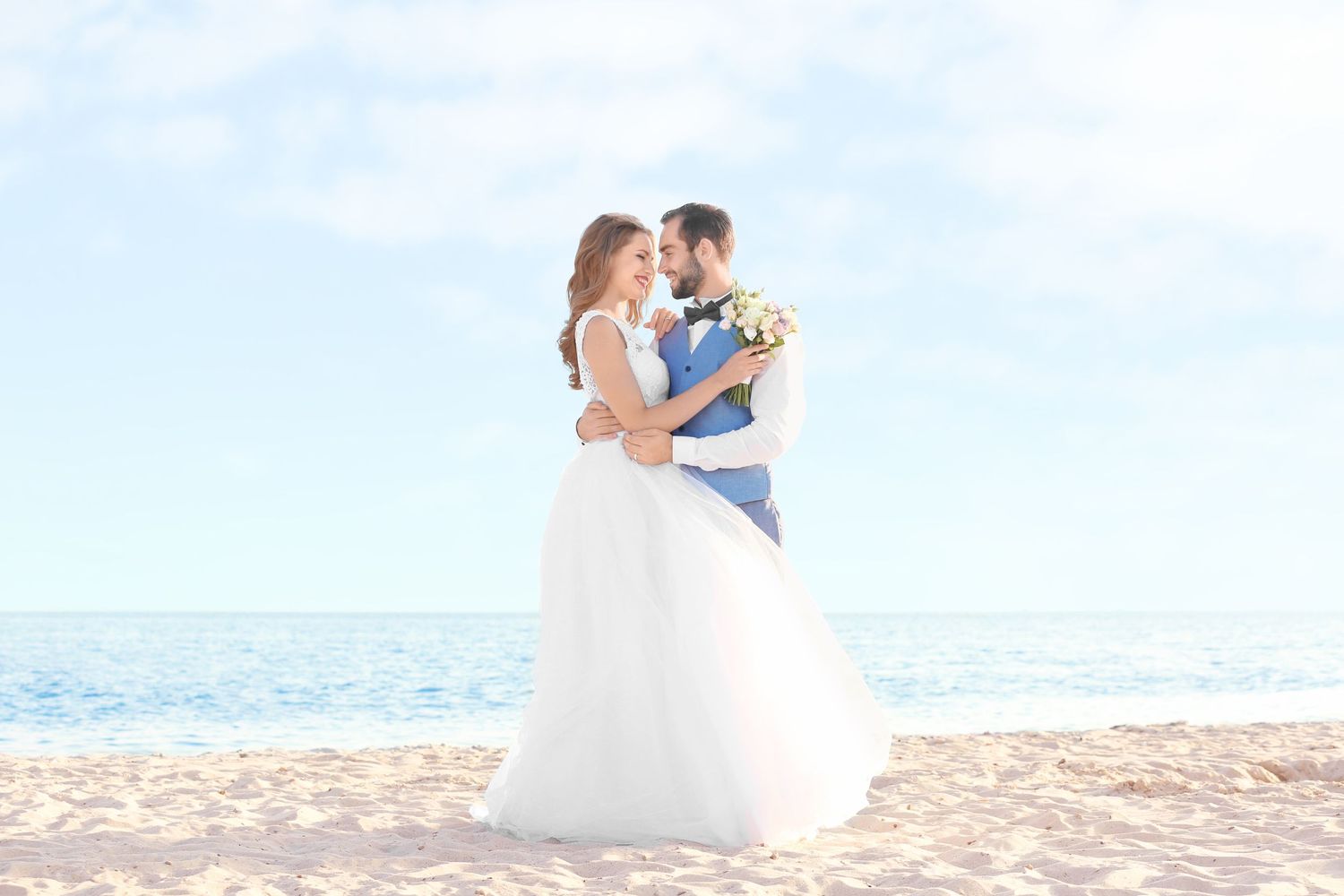 Couple in wedding attire embracing on sunny beach with ocean backdrop.