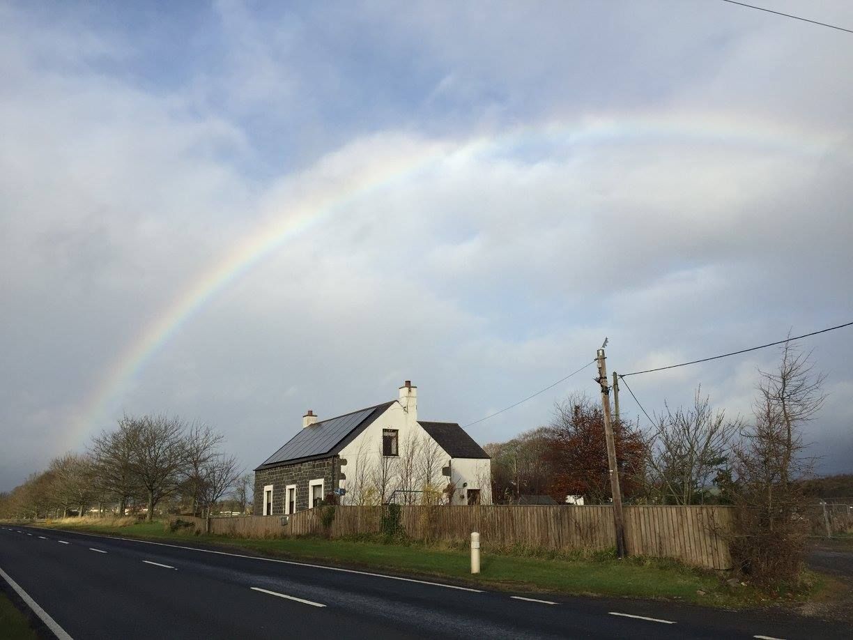 Beautiful rainbow arches over a rural house beside country road on cloudy day.