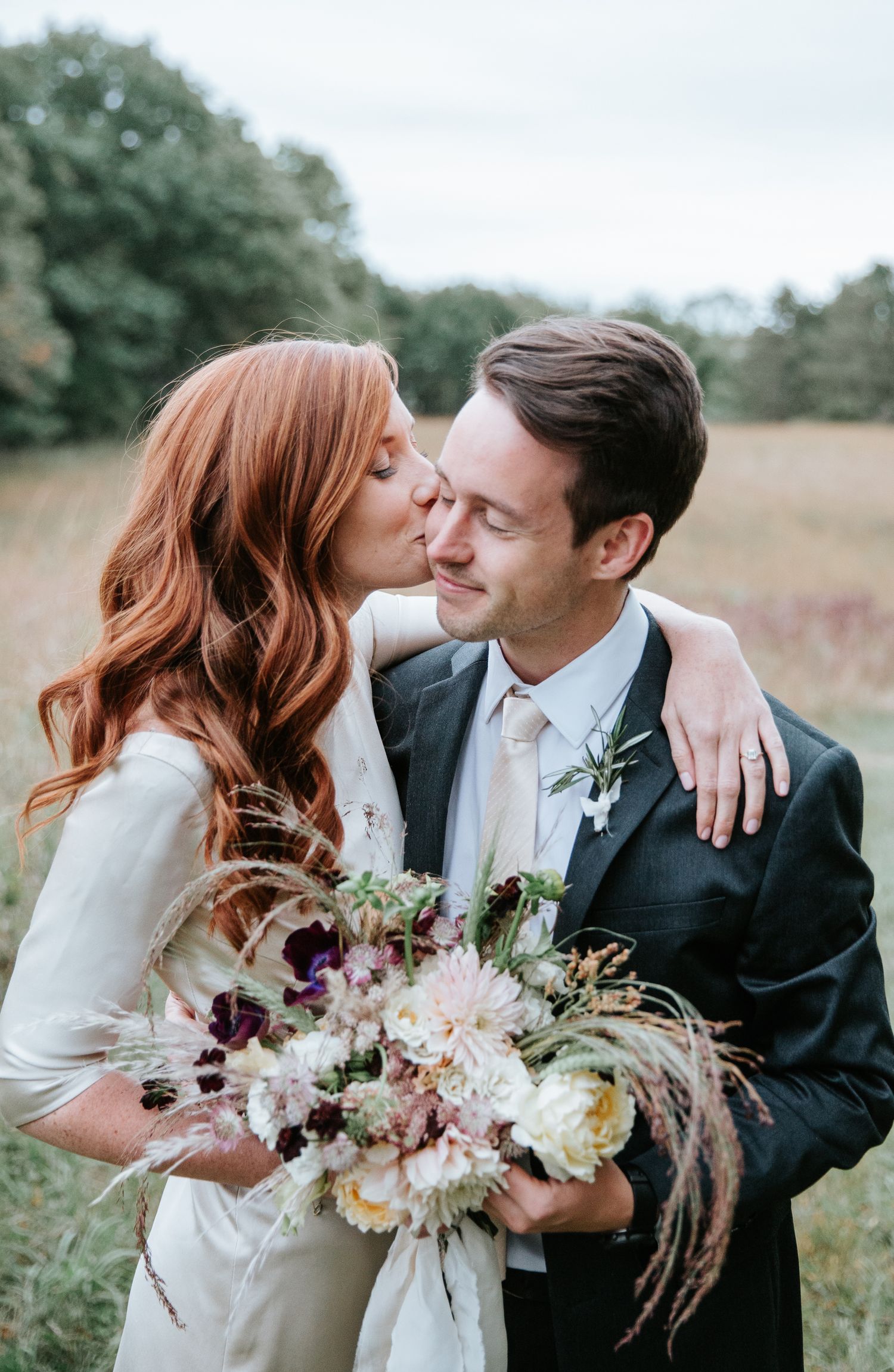 redhead bride kisses brunette groom in meadow at their outdoor couple shoot