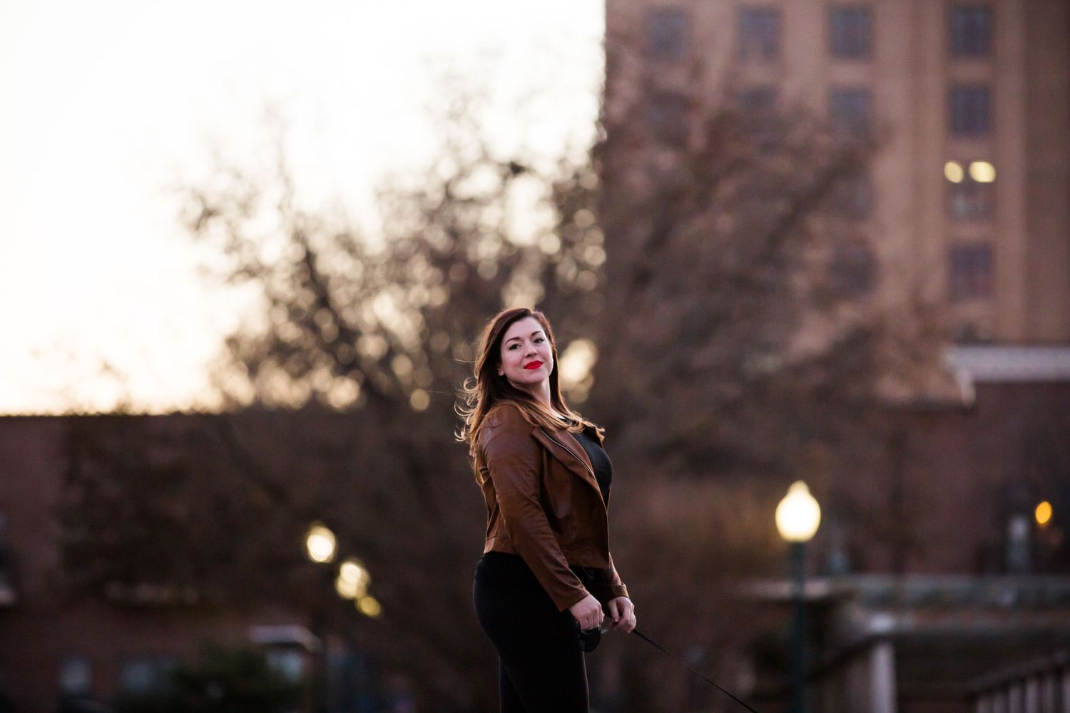 Woman in brown jacket poses at sunset with urban buildings in background.
