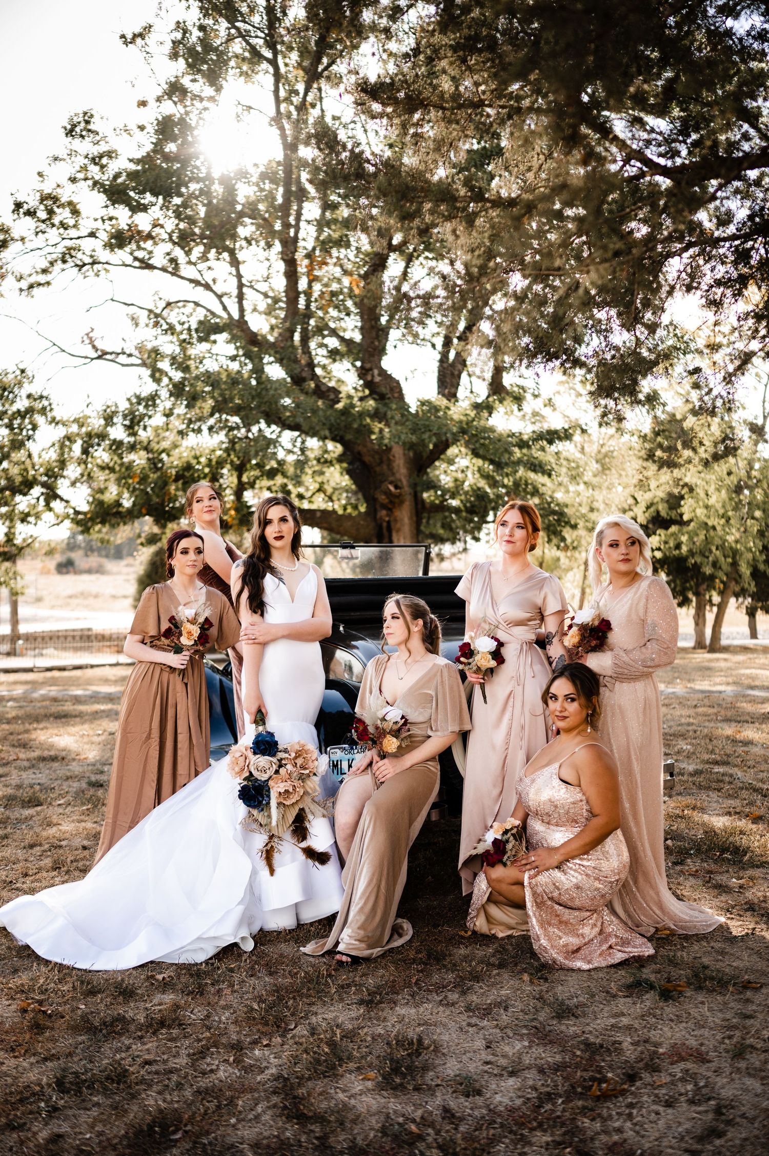 Bridal party in neutral tones poses with vintage car under sunlit trees.
