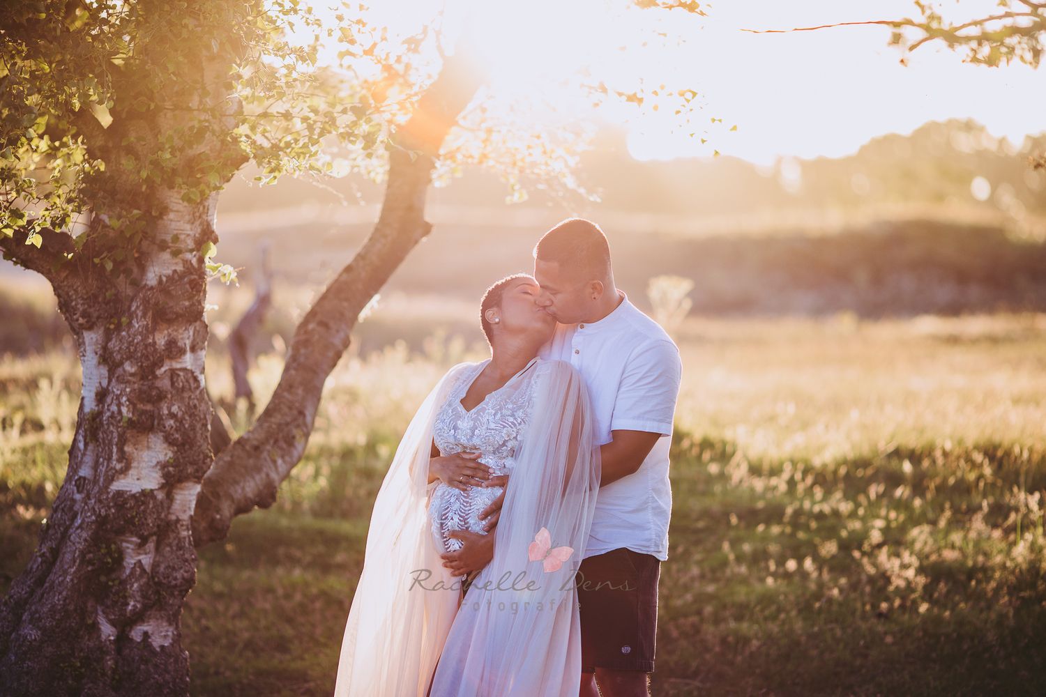 Romantic sunset maternity photo of couple embracing in white outfits under trees.
