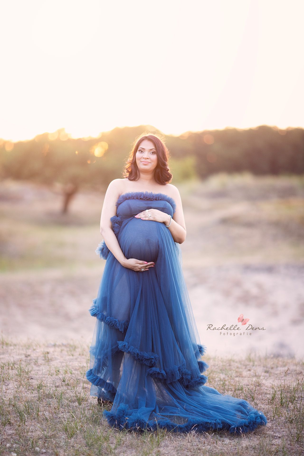 Dramatic maternity portrait in flowing blue tulle gown at golden sunset.