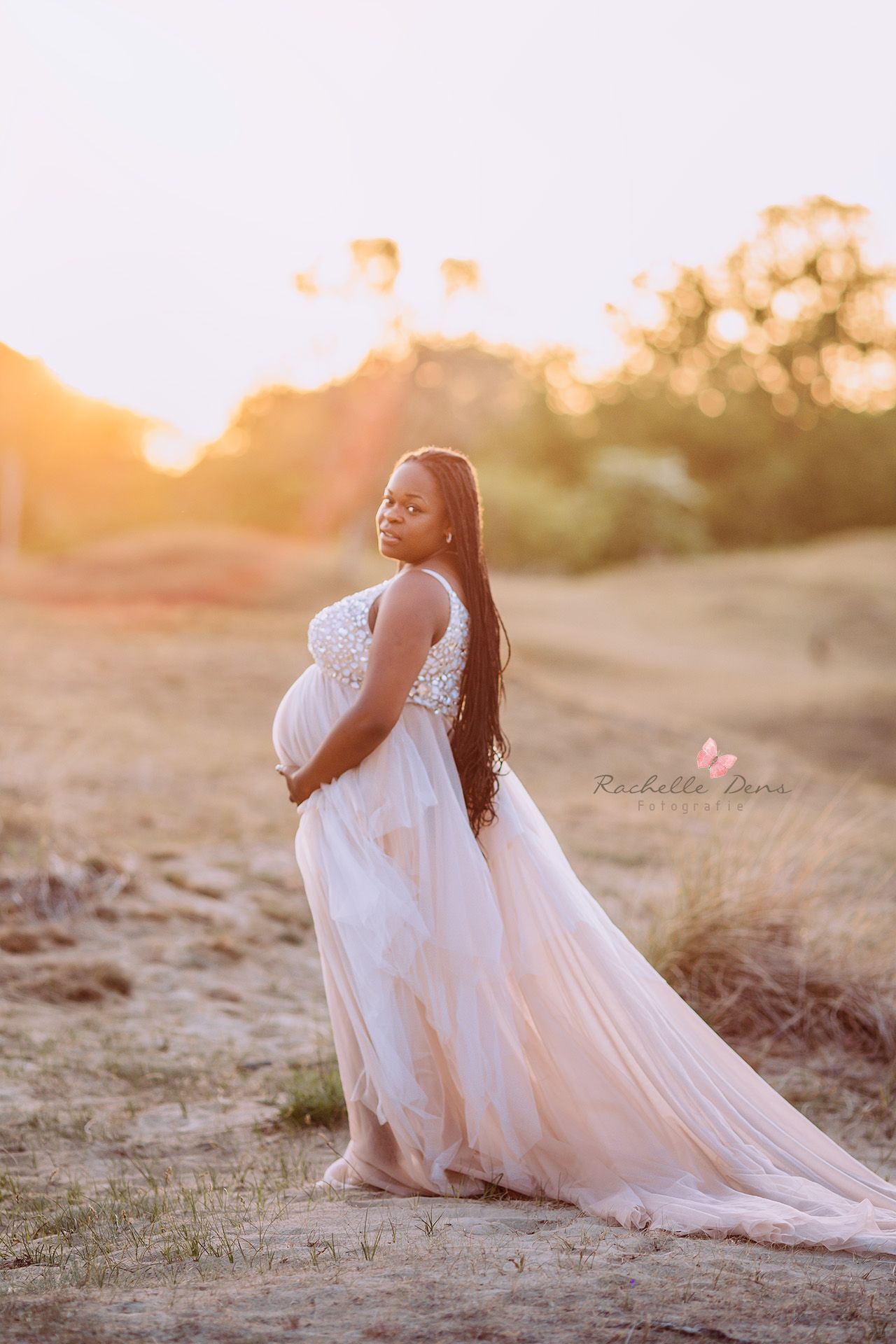 Elegant maternity photo in white lace gown during golden hour outdoors.