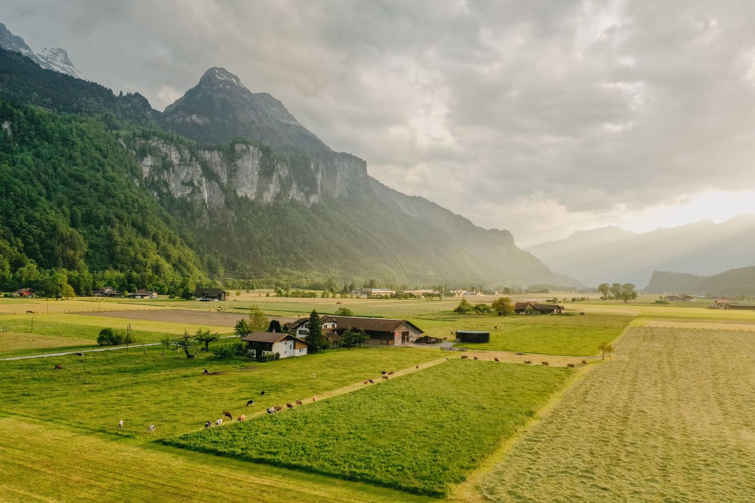 A scenic mountain landscape with green meadows and farmland nestled beneath dramatic rocky peaks in soft sunlight.