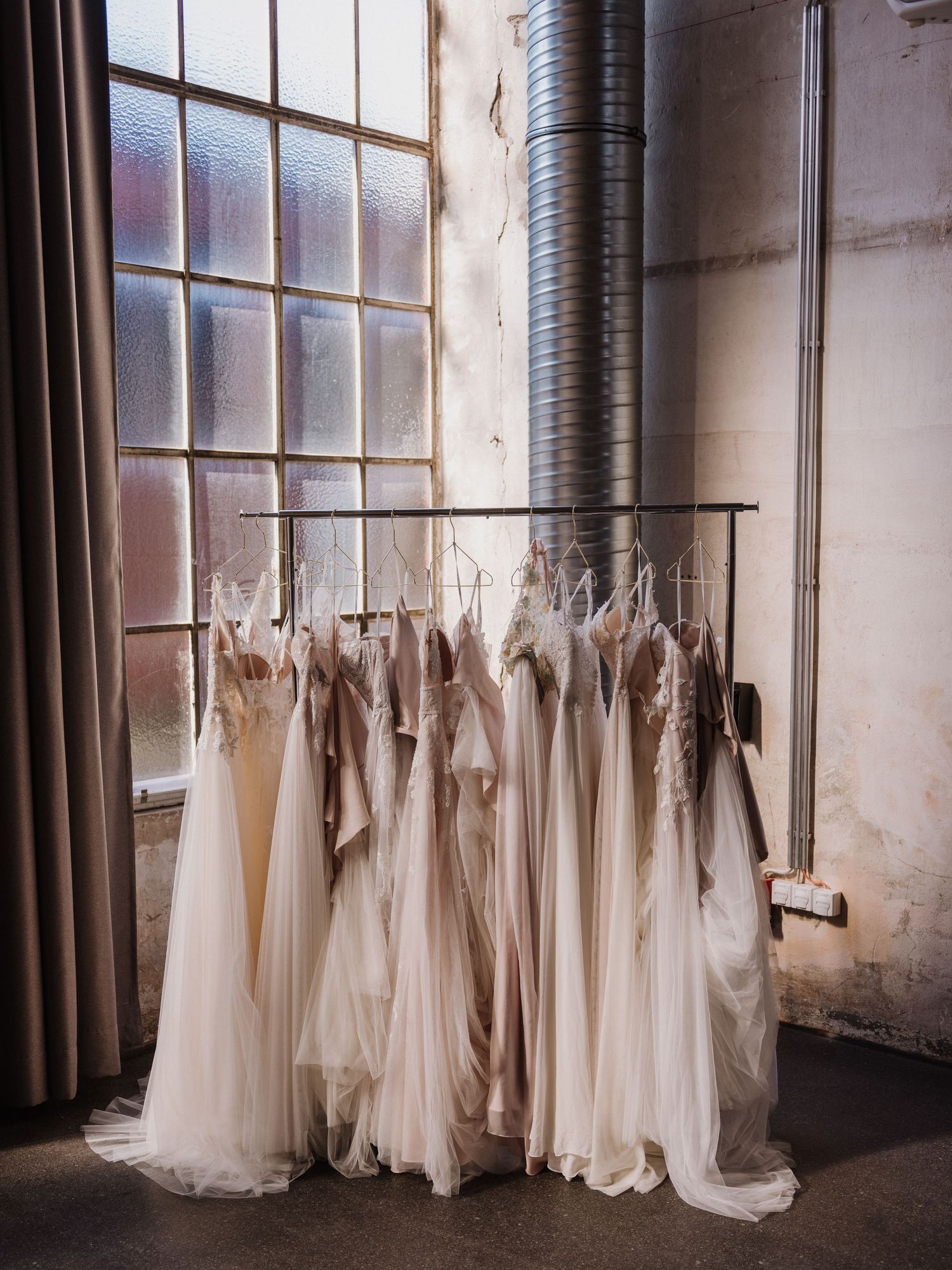 Row of white wedding dresses hanging on rack near industrial windows.