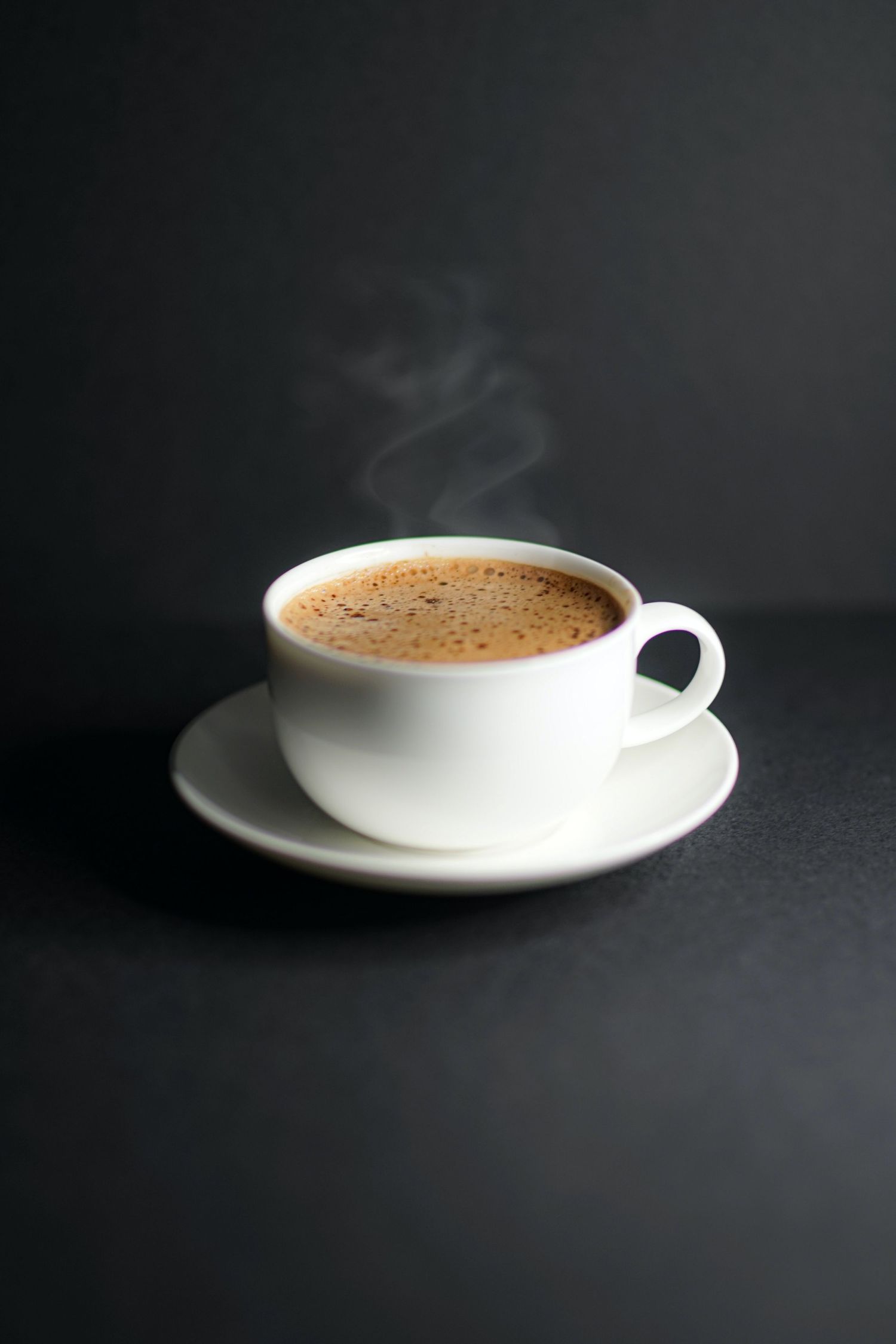 A steaming cup of fresh coffee in a white porcelain cup and saucer against a dark background.