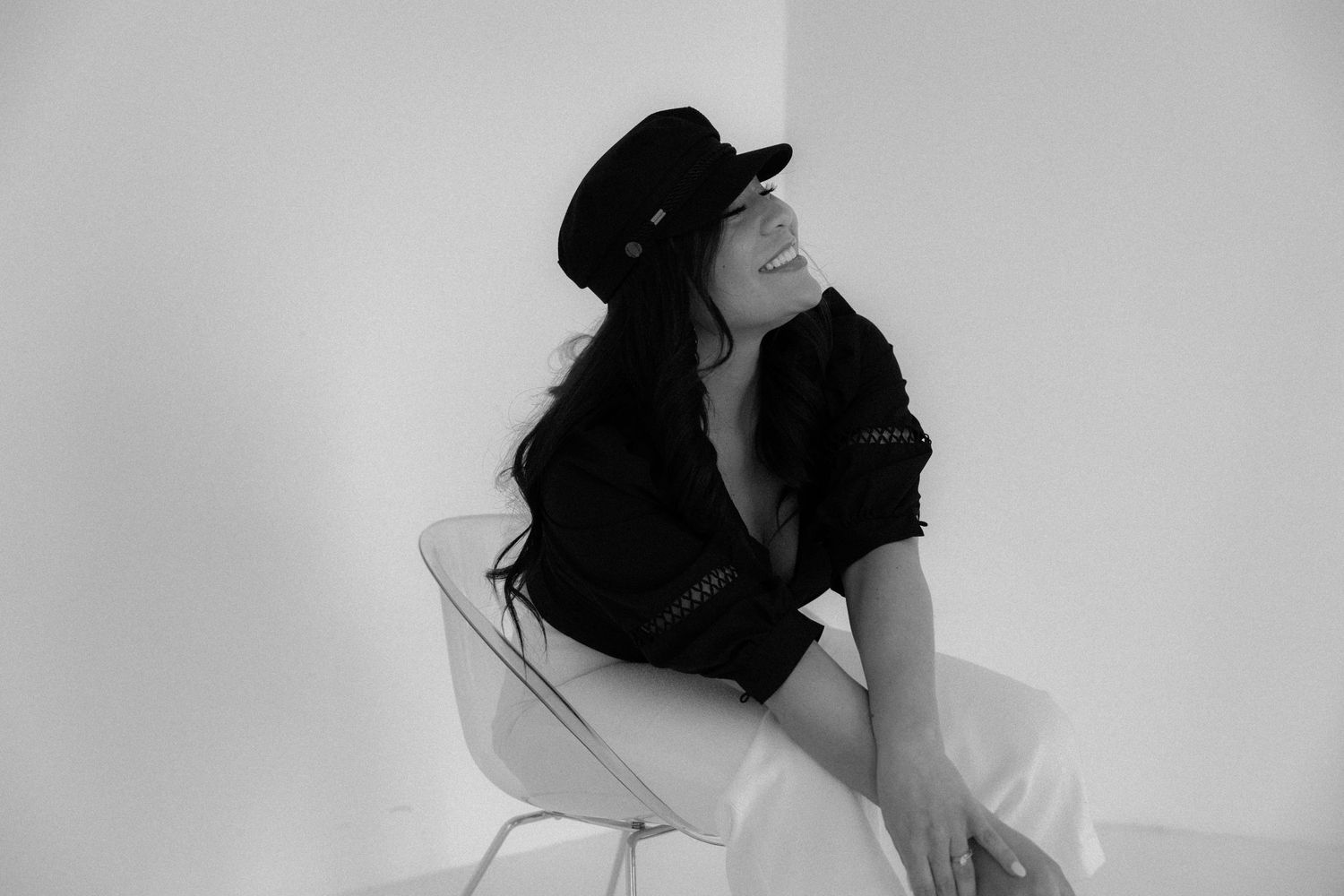 Black and white portrait of smiling woman in baker boy cap seated in white chair.