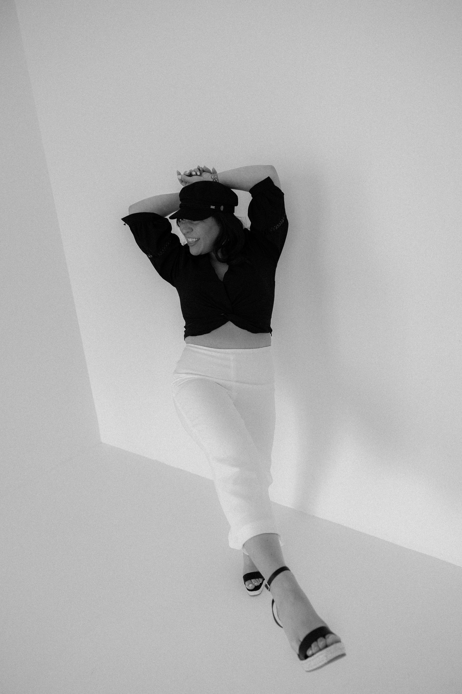 Black and white photo of woman in dark cap and white skirt leaning against white wall.