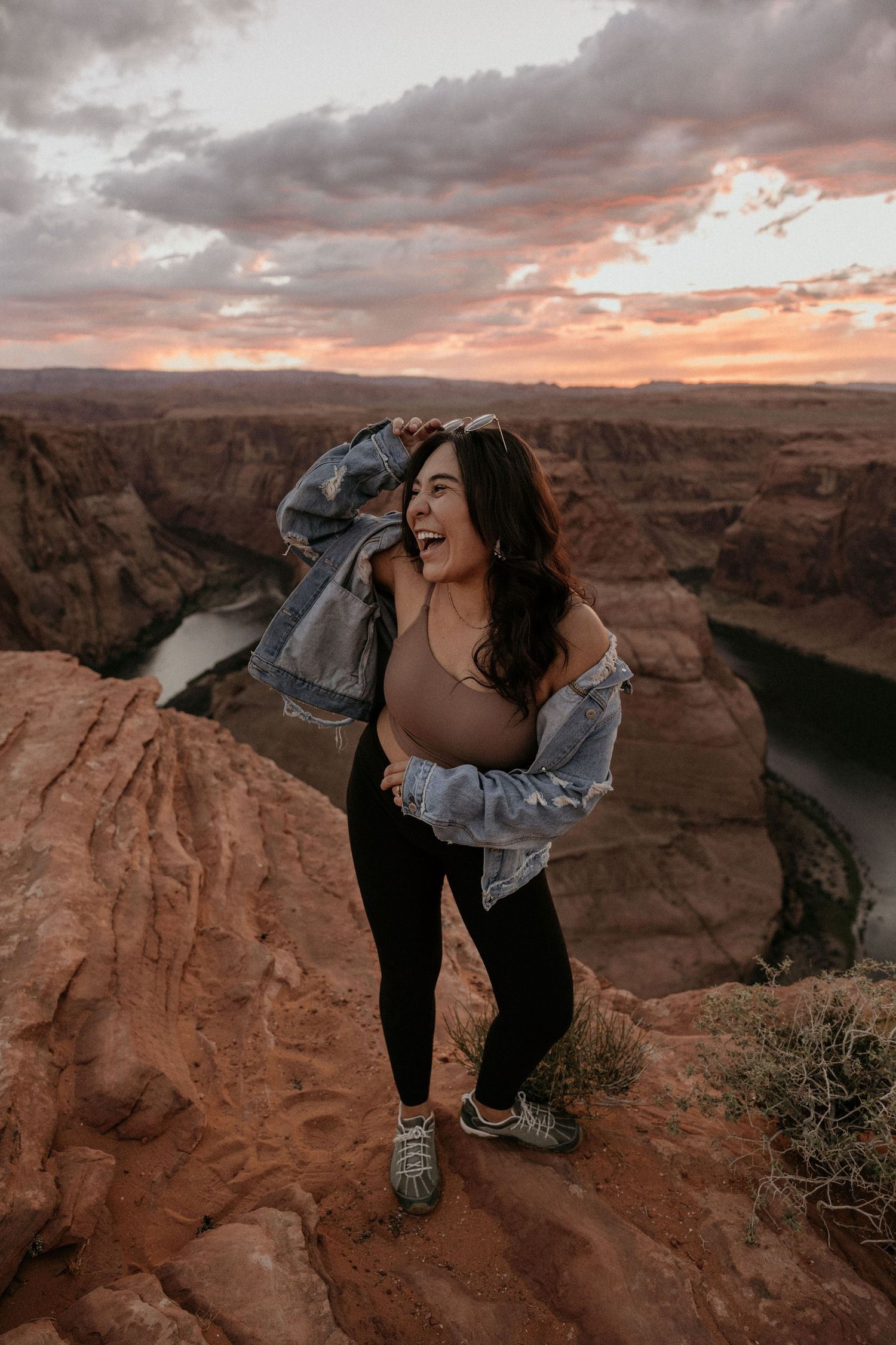 Woman laughing at sunset at Horseshoe Bend overlook wearing denim jacket and black leggings.