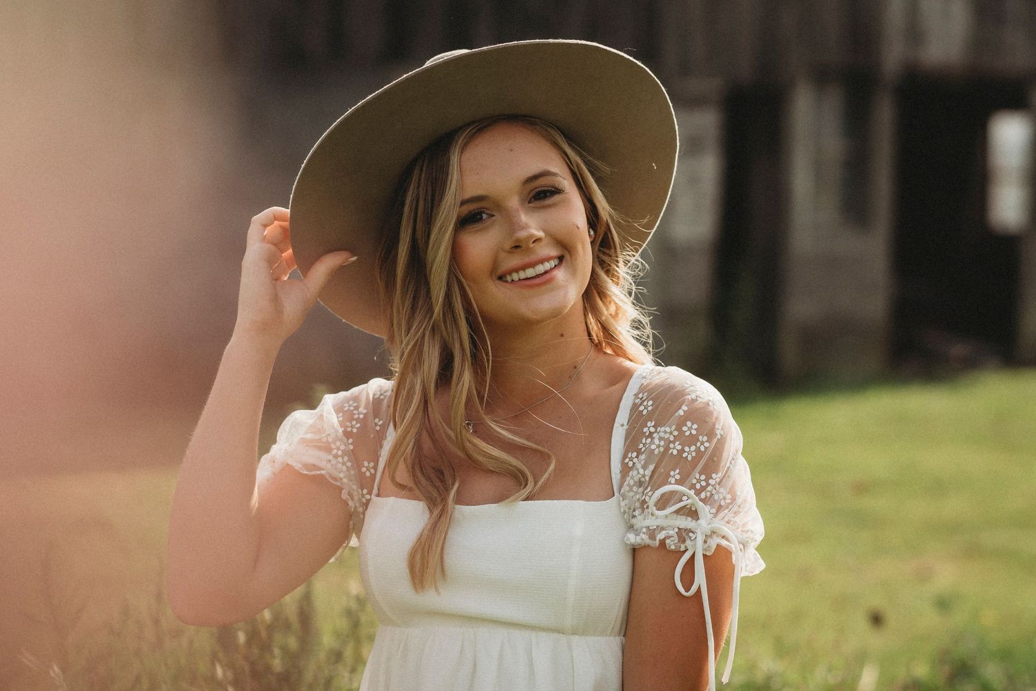 Someone in a white dress and tan hat poses in warm sunlight on a summer evening.