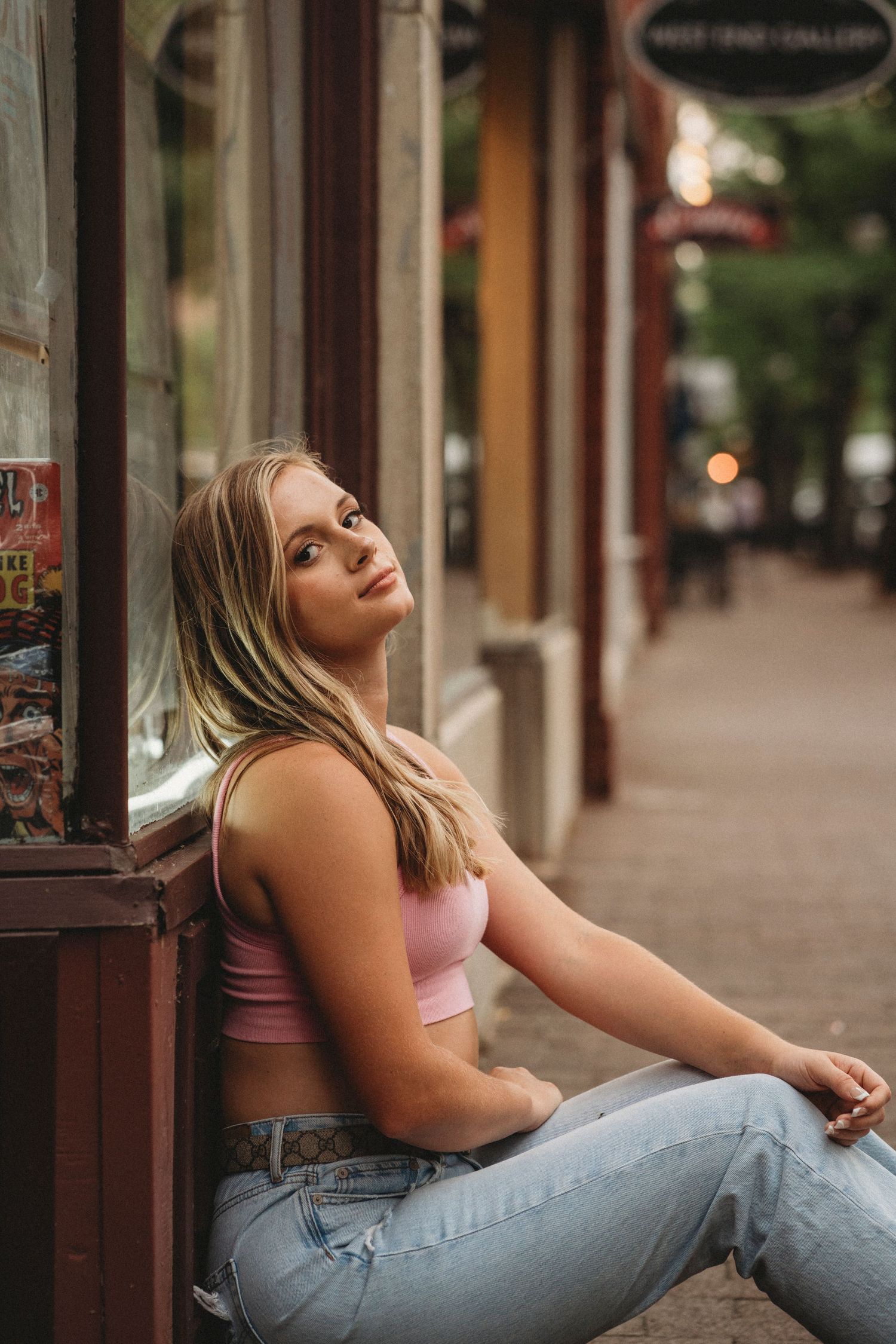 A person in a pink crop top and jeans sits casually against a storefront wall on a city sidewalk.