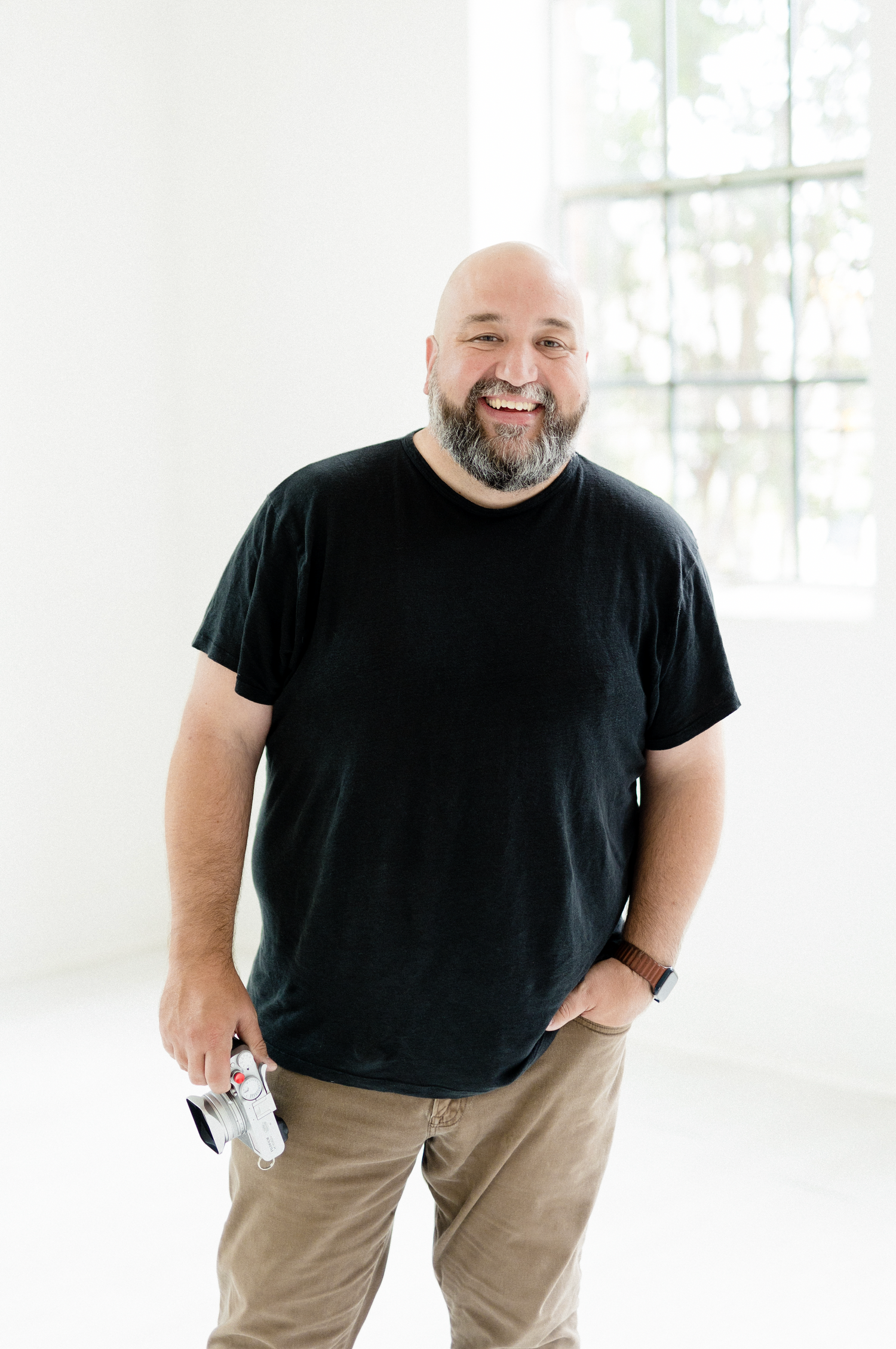 Photographer holding camera while standing in bright studio wearing black t-shirt and khaki pants.