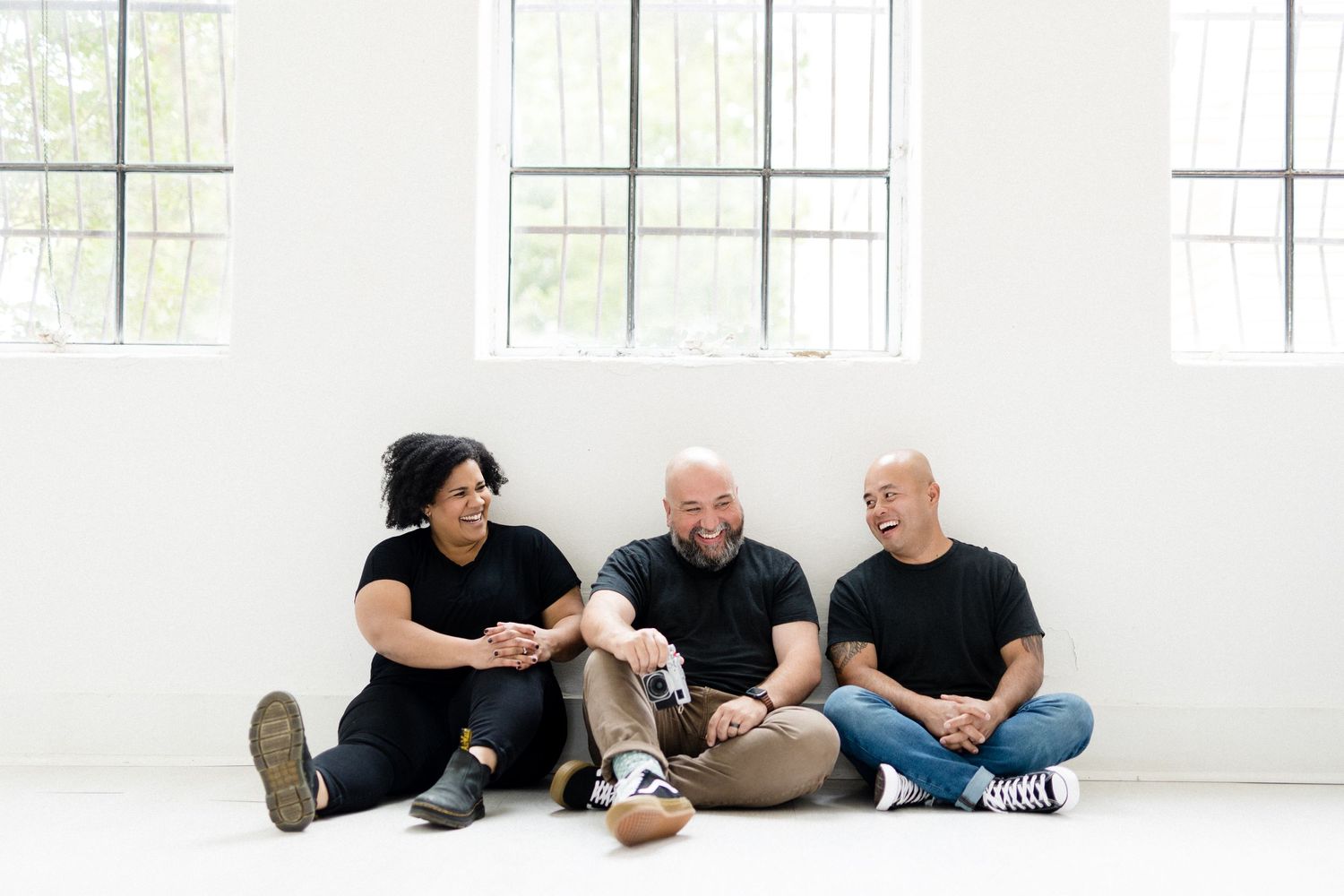 Three people relaxing casually on floor of bright white room with windows.