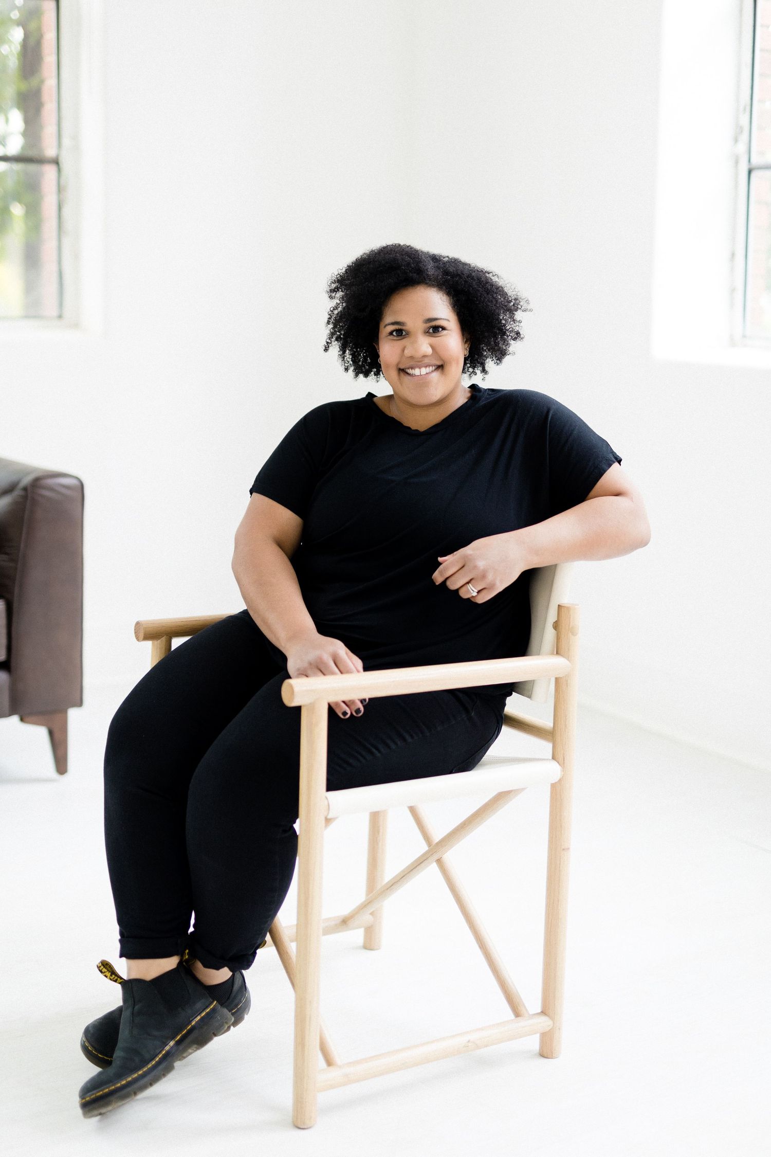 Person in black outfit sitting comfortably in director's chair in bright studio setting.