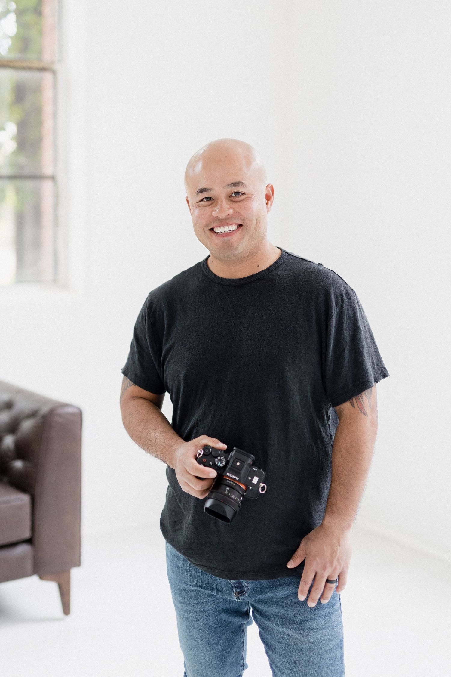 Photographer in black t-shirt holding camera while standing in bright studio space.