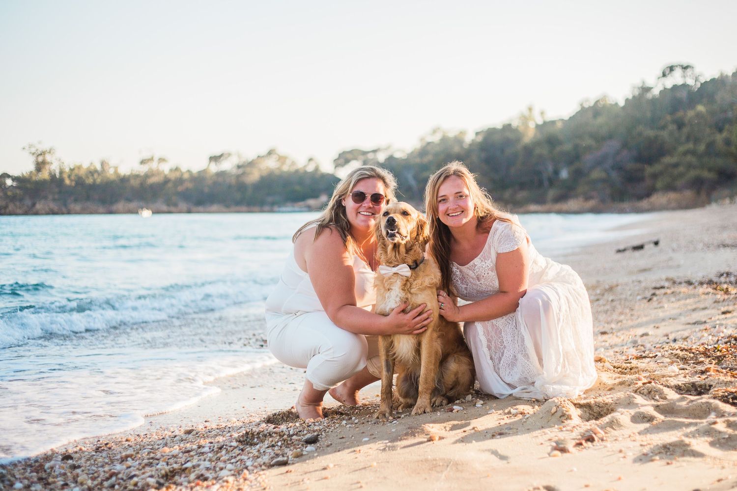 two women in wedding attire on a sandy beach with their golden retriever dog in a bow tie