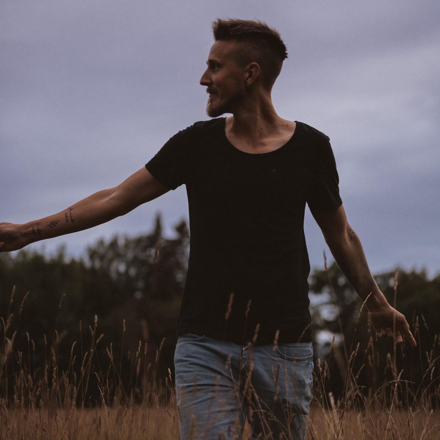 Silhouette of person in black shirt and jeans standing in tall grass at dusk.