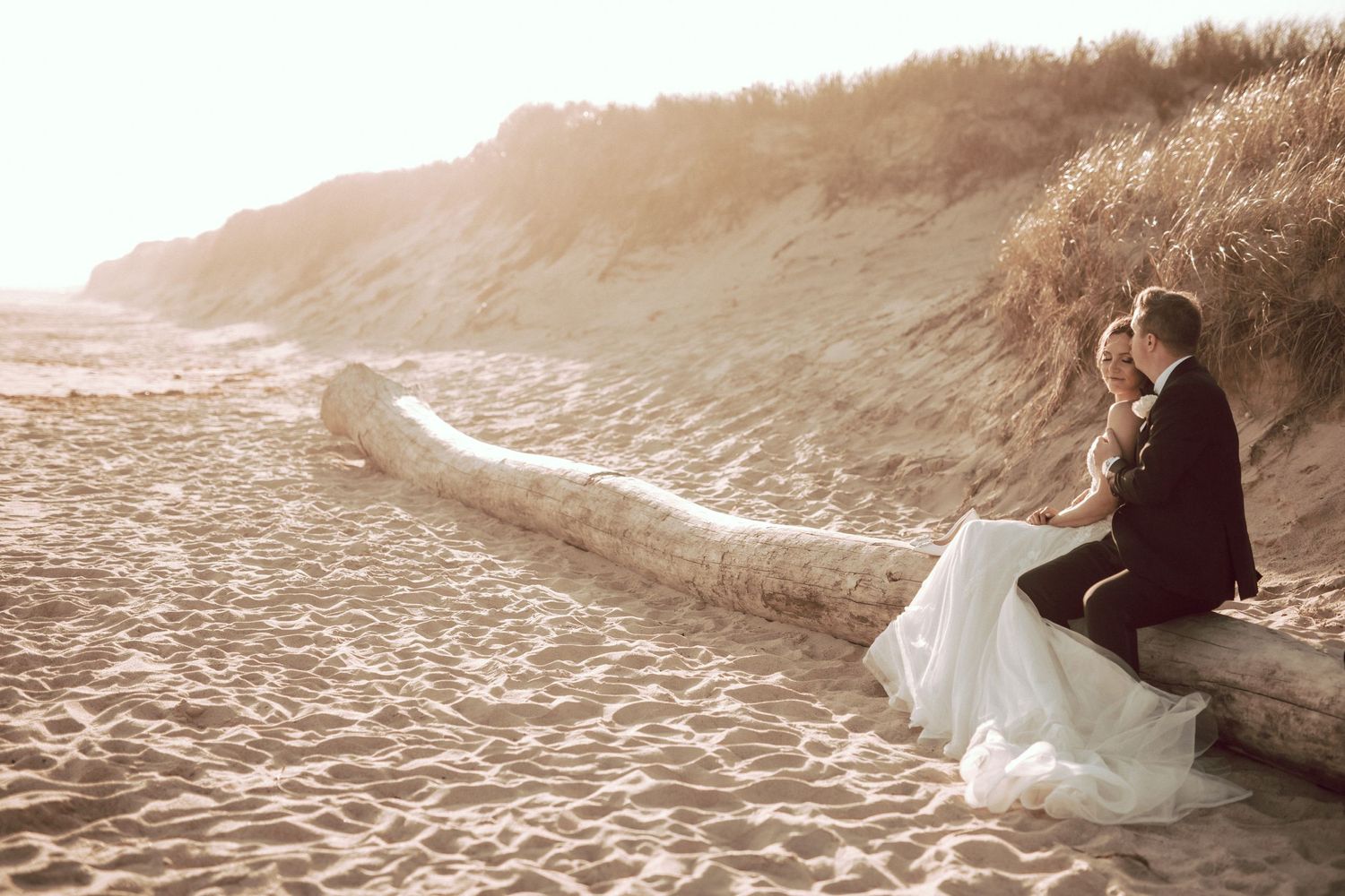 A couple of newlyweds taking in the view on the beach in Montauk sitting on a piece of drift wood reminiscing about their day before heading to evening celebration at 360 East at Montauk Downs