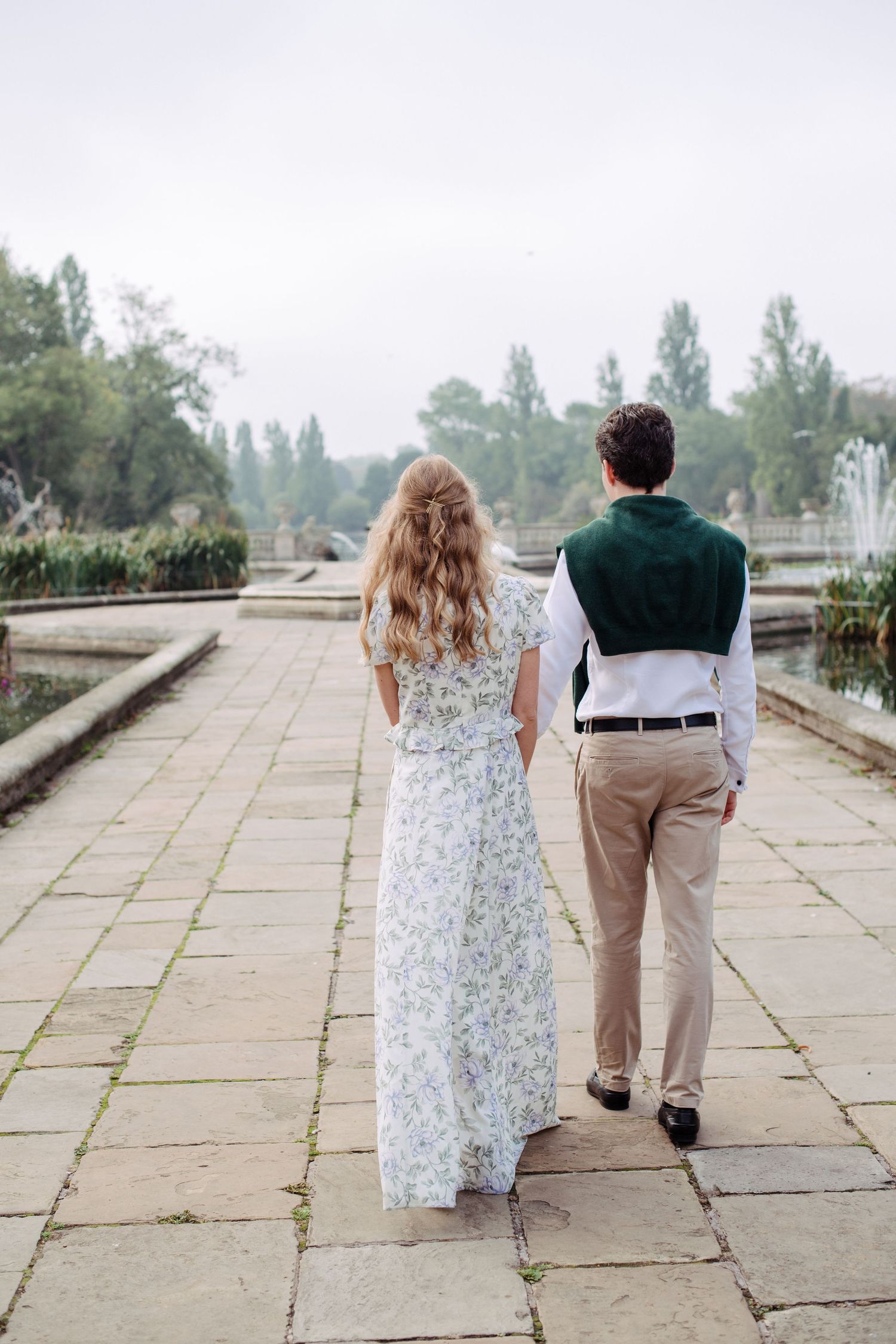 A couple walks hand in hand along a scenic stone path by the water, with the woman wearing a floral dress.