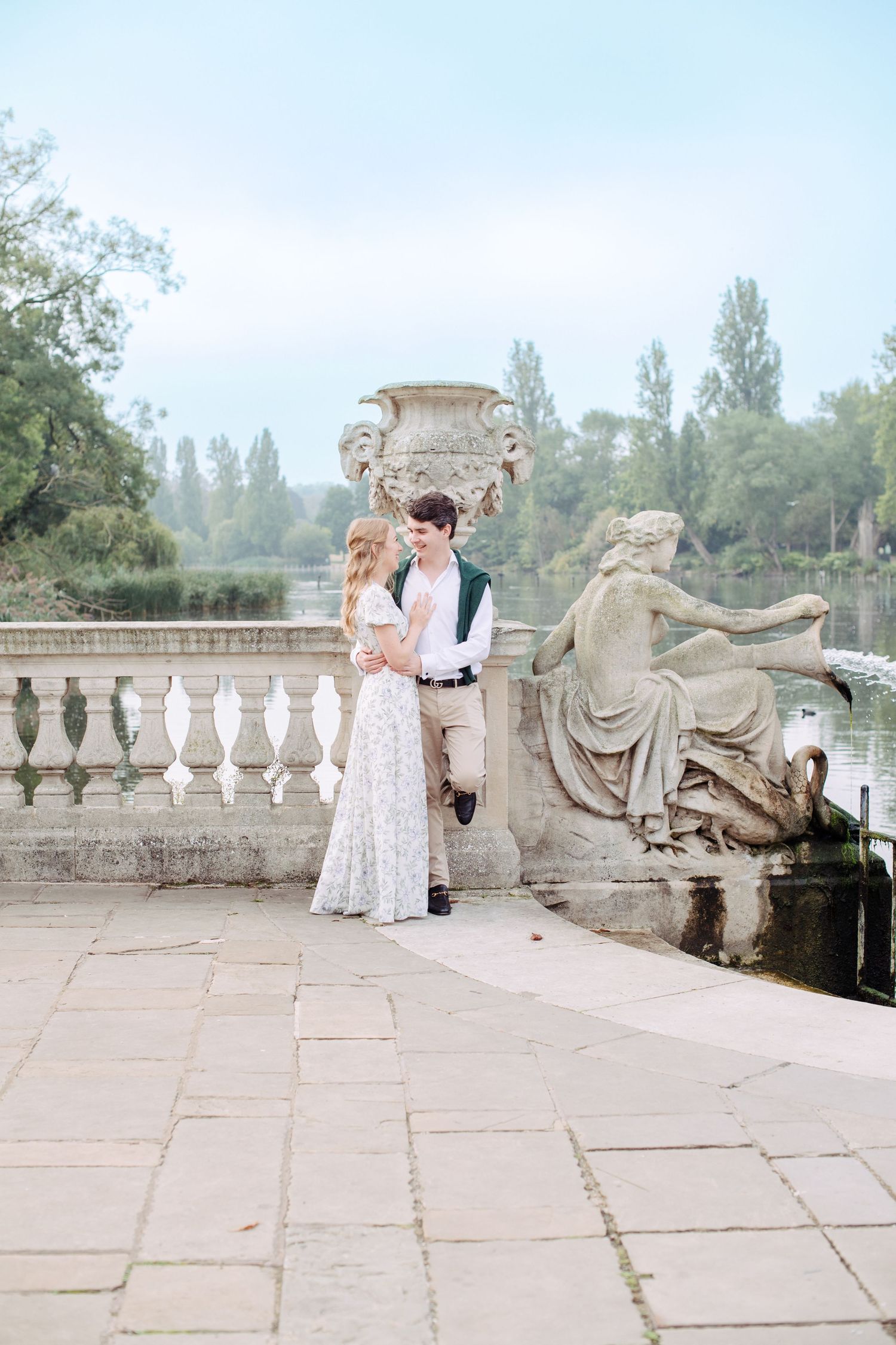 A couple stands on an ornate stone terrace with classical statuary and architectural details in a romantic garden setting. Enjoying London
