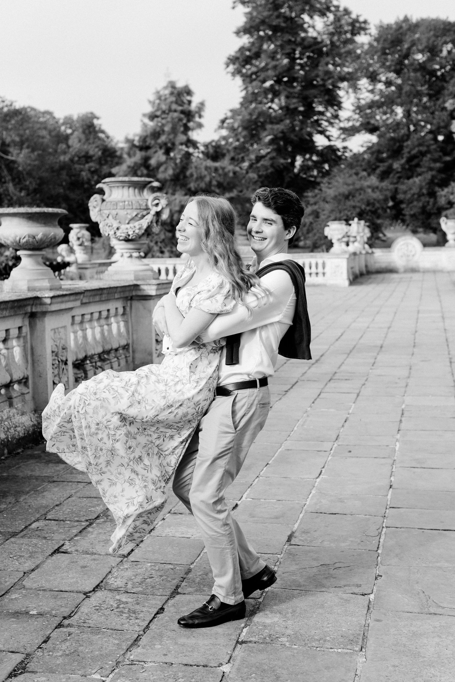 A romantic couple dances together on a stone terrace with decorative urns and lush gardens in the background on a sunny day in London.