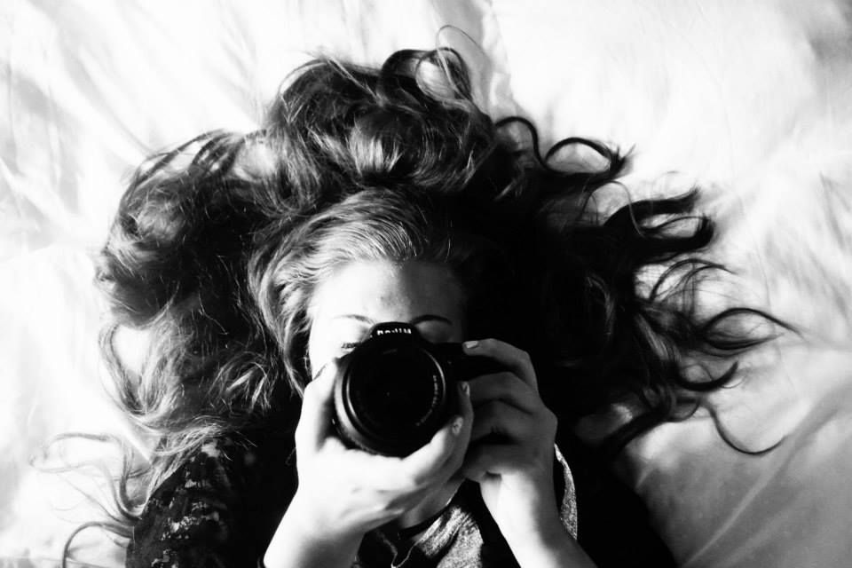 A black and white photo of curly hair and a camera lens pointed at the viewer.
