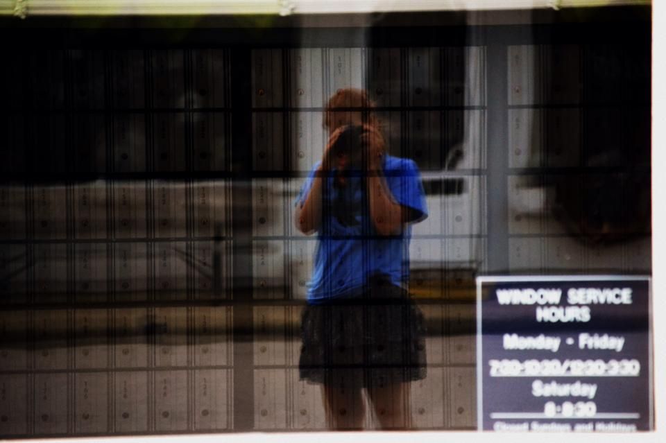 A blurry reflection of a photographer in a window with business hours posted.