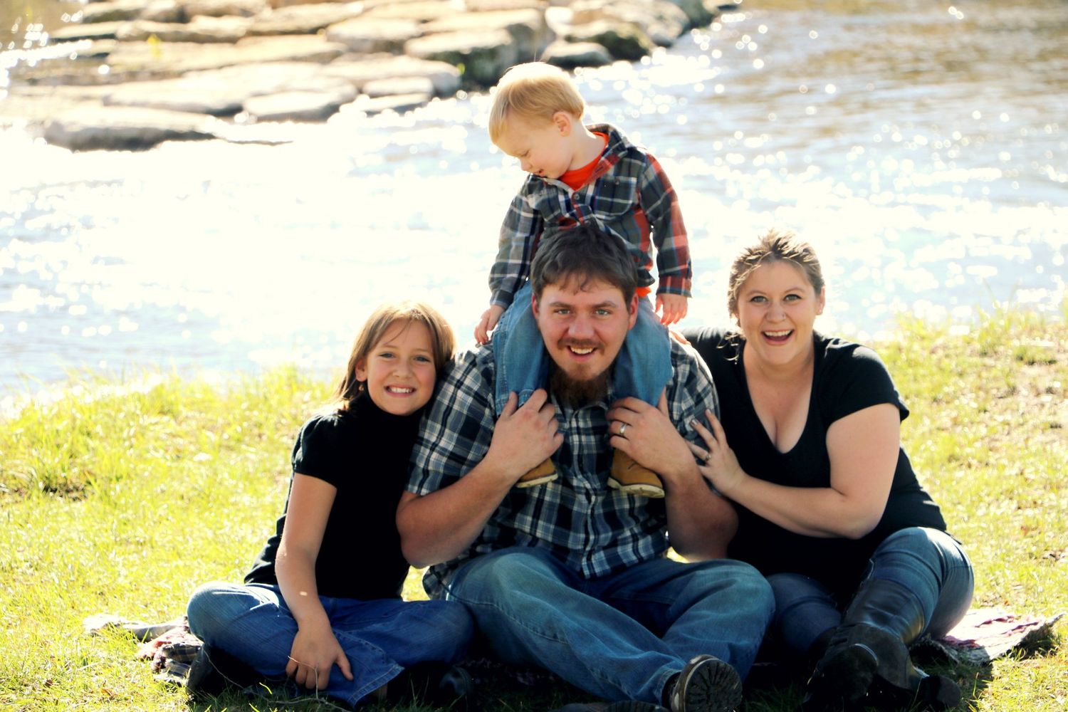 Family group sitting together on grass near water's edge enjoying a sunny outdoor day.
