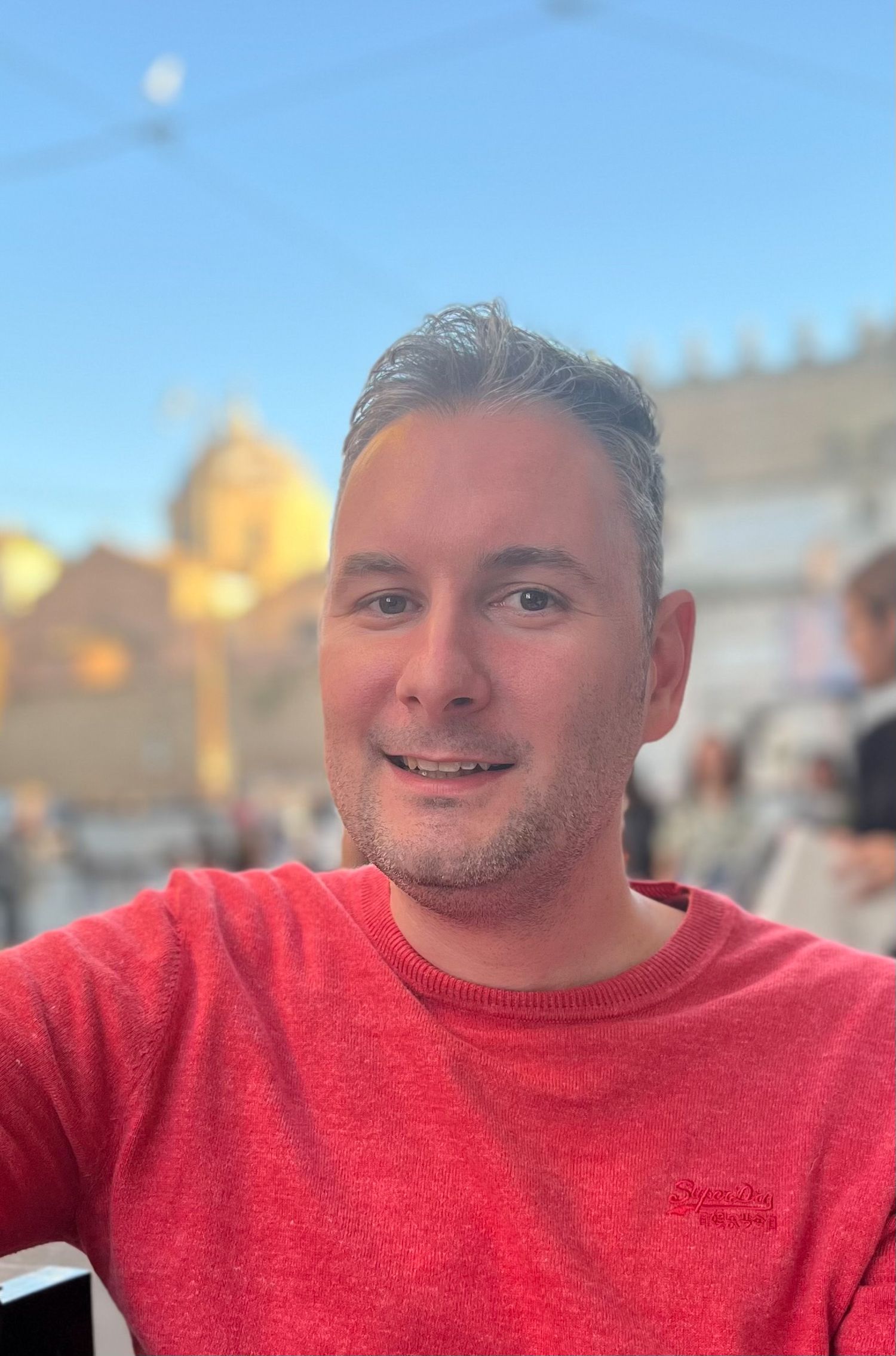 A person in a red sweater smiles while sitting outdoors with buildings in the background.