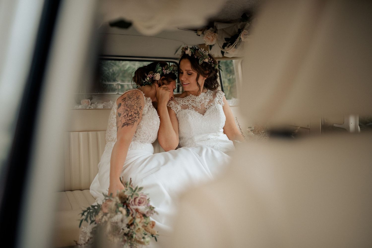 Two brides share an intimate moment in the back of a vintage wedding car with soft light and floral bouquets.