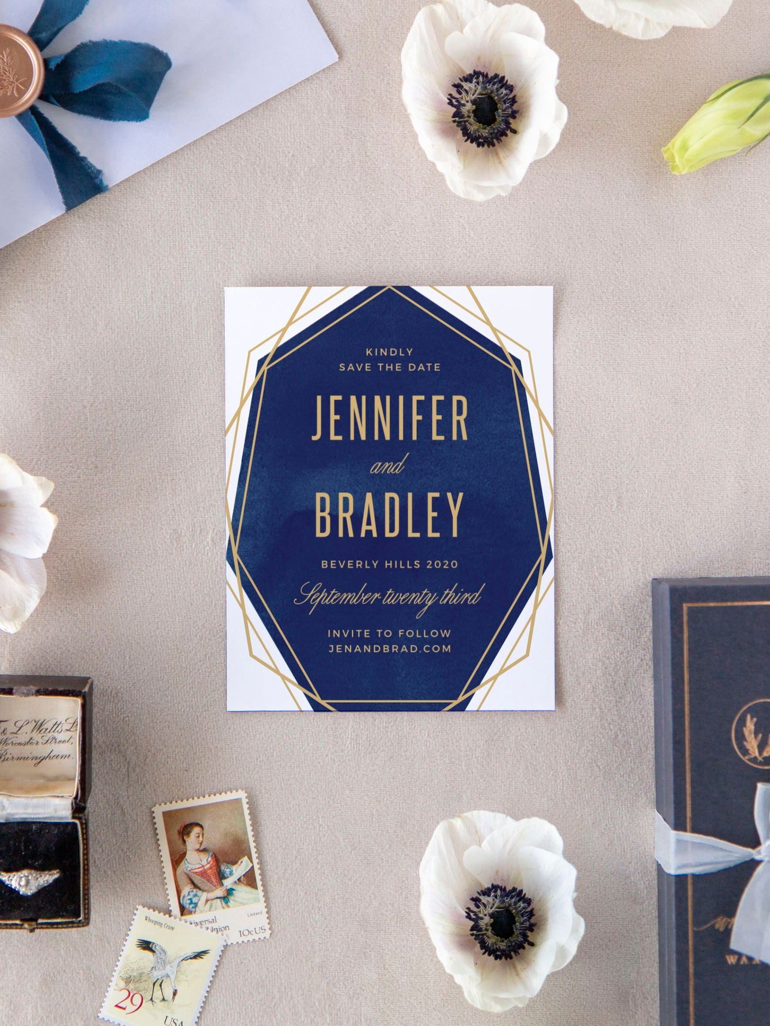 Navy and gold geometric wedding invitation with white flowers and rings on a neutral background.