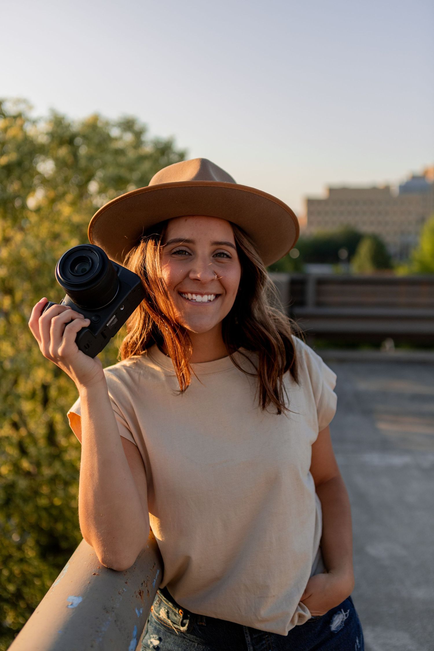 Photographer in tan hat holds camera while smiling during golden hour photoshoot.