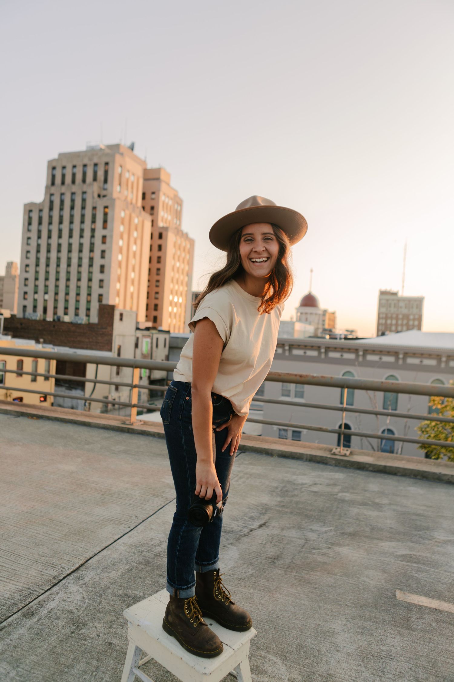 Person in casual attire and hat posing playfully on an urban rooftop with city buildings in background.