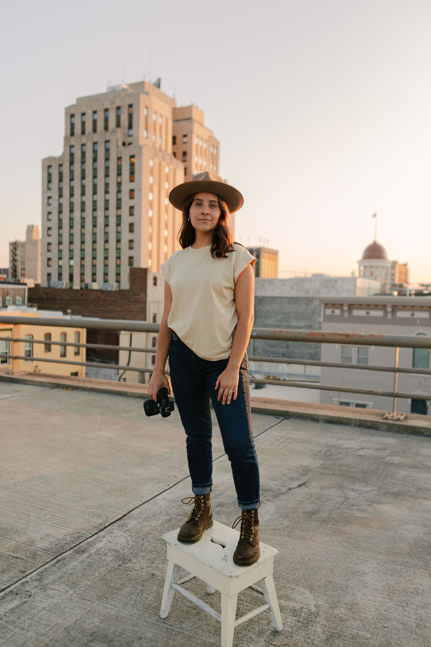 A person in jeans and boots poses on a white stool on a rooftop at sunset with city buildings in the background.