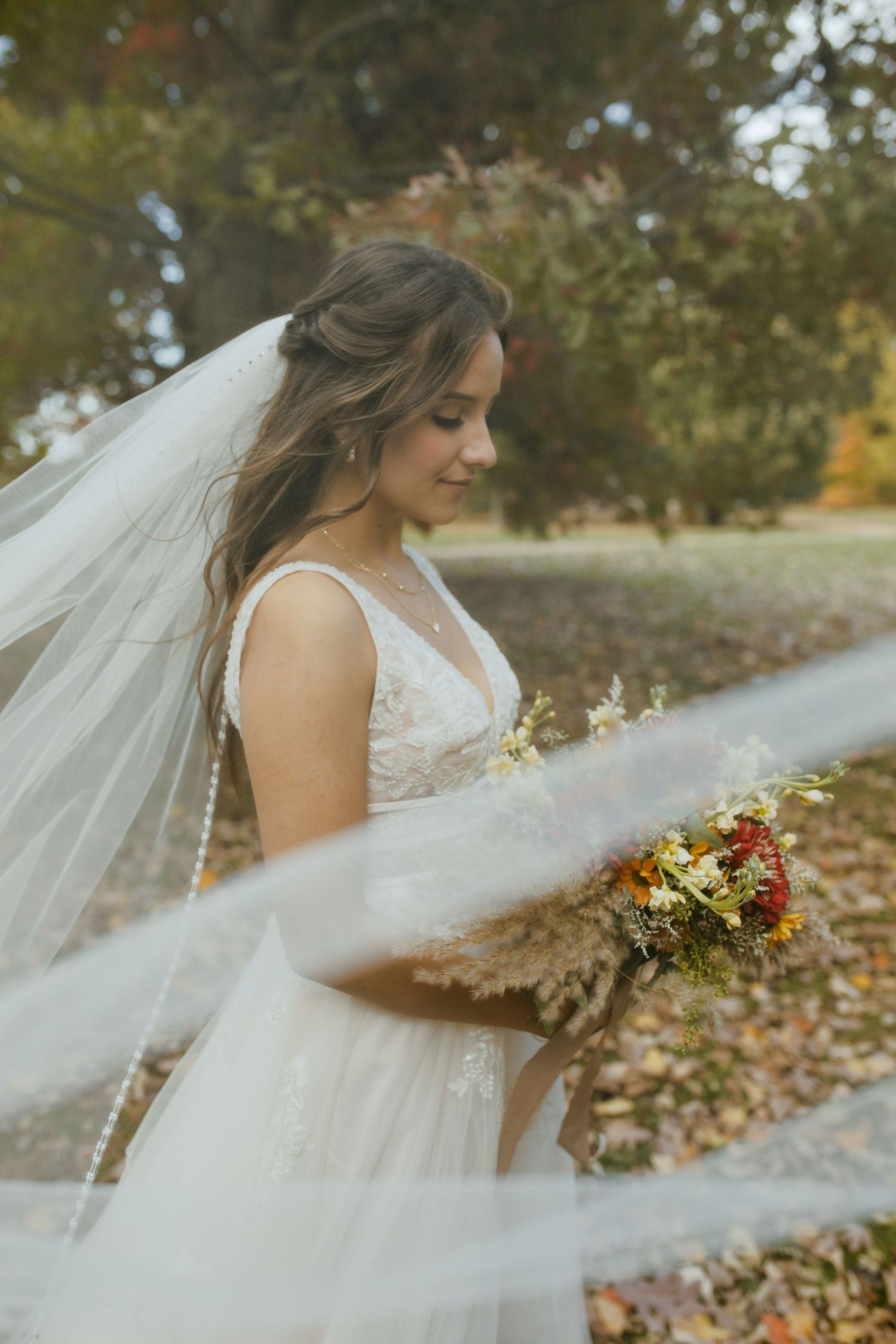 A bride in a flowing white dress holds a colorful autumn bouquet while her veil catches the breeze.