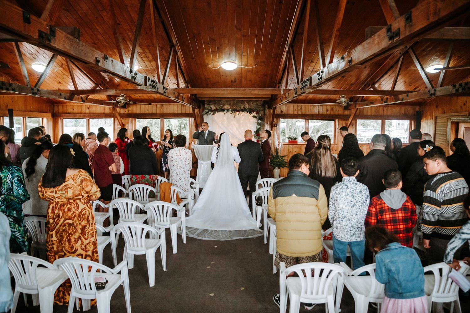 bride and groom standing at the end of the isle with the officiant, in a wooden building with many windows.