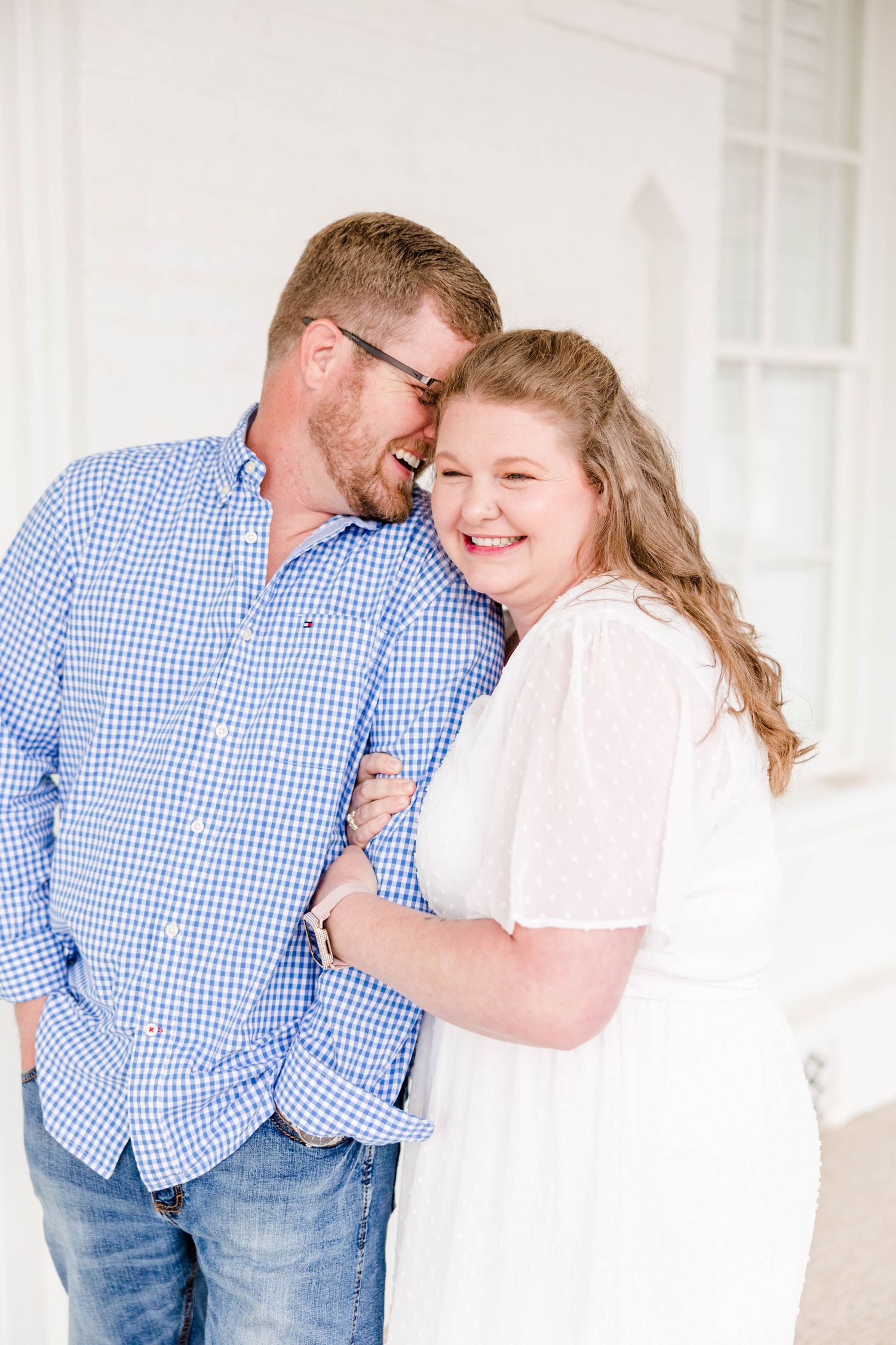 A couple shares joyful laughter while embracing in a bright white hallway.